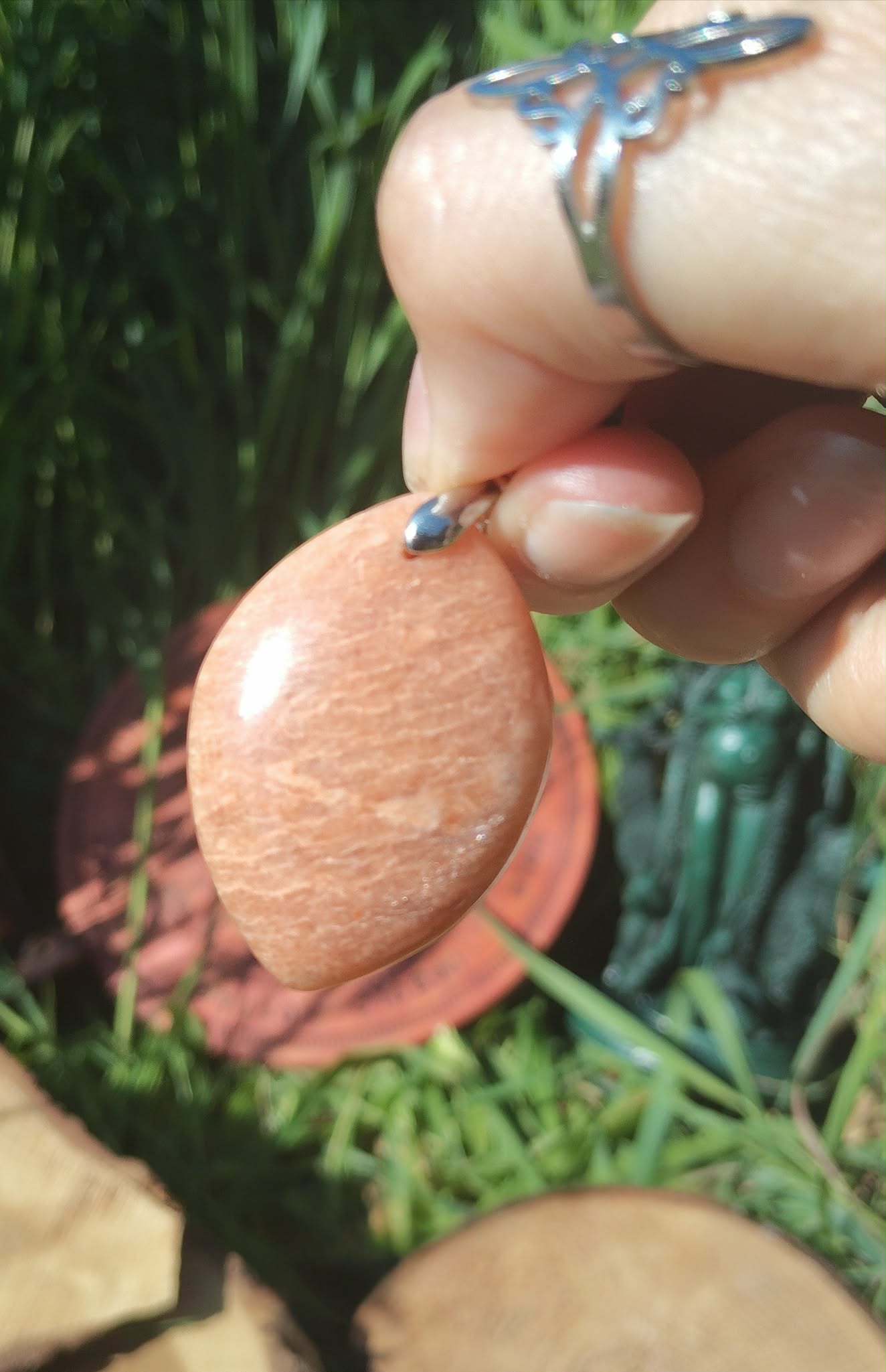 Pendentif goutte en pierre de soleil tenu en main sur feuilles vertes, pierre naturelle associée à la spontanéité, au courage paisible et à la chaleur du cœur.