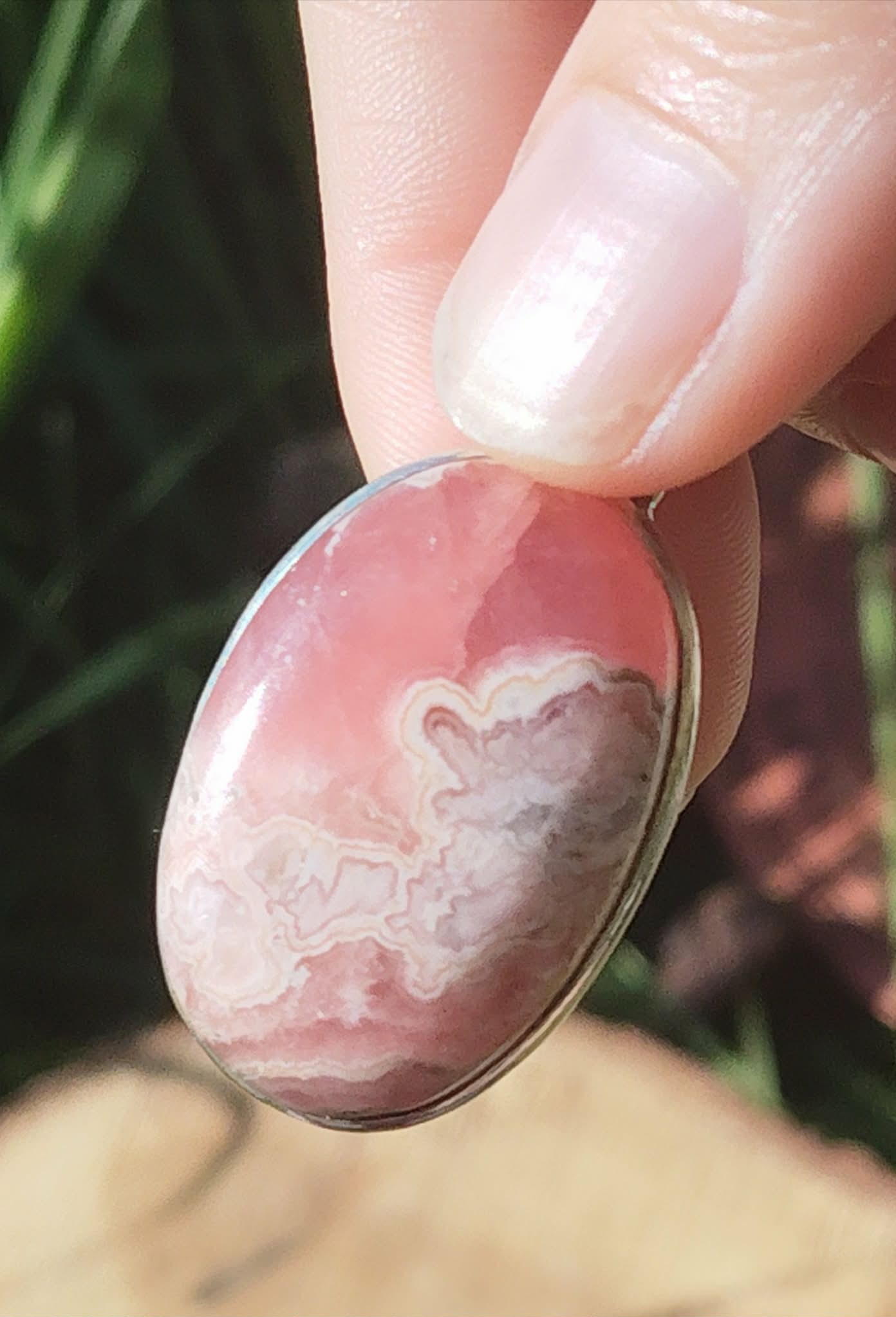 Gros plan sur pendentif rhodochrosite ovale rose marbré avec monture argent 925, tenu entre les doigts devant fond végétal et champignon, bijou en pierre naturelle associé à la sensibilité apaisée, à la douceur et à l’équilibre du cœur.