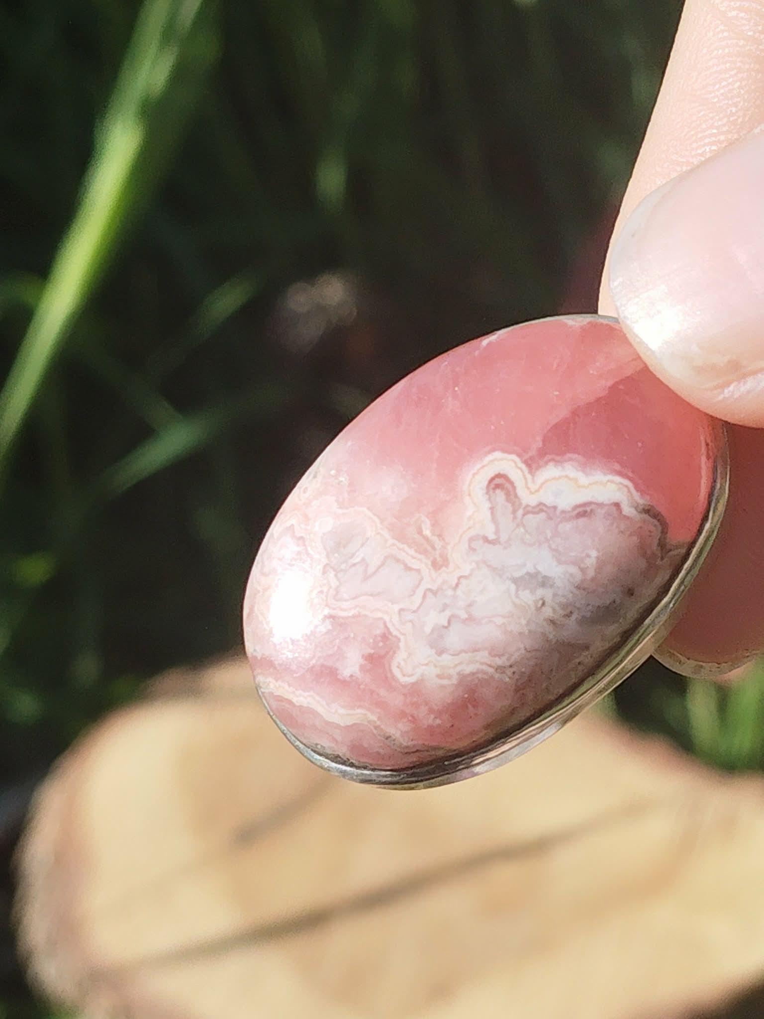 Gros plan sur pendentif rhodochrosite ovale rose marbré serti argenté, tenu entre les doigts avec fond végétal et bois, bijou en pierre naturelle lié au réconfort, à la douceur du cœur et à la stabilité intérieure.