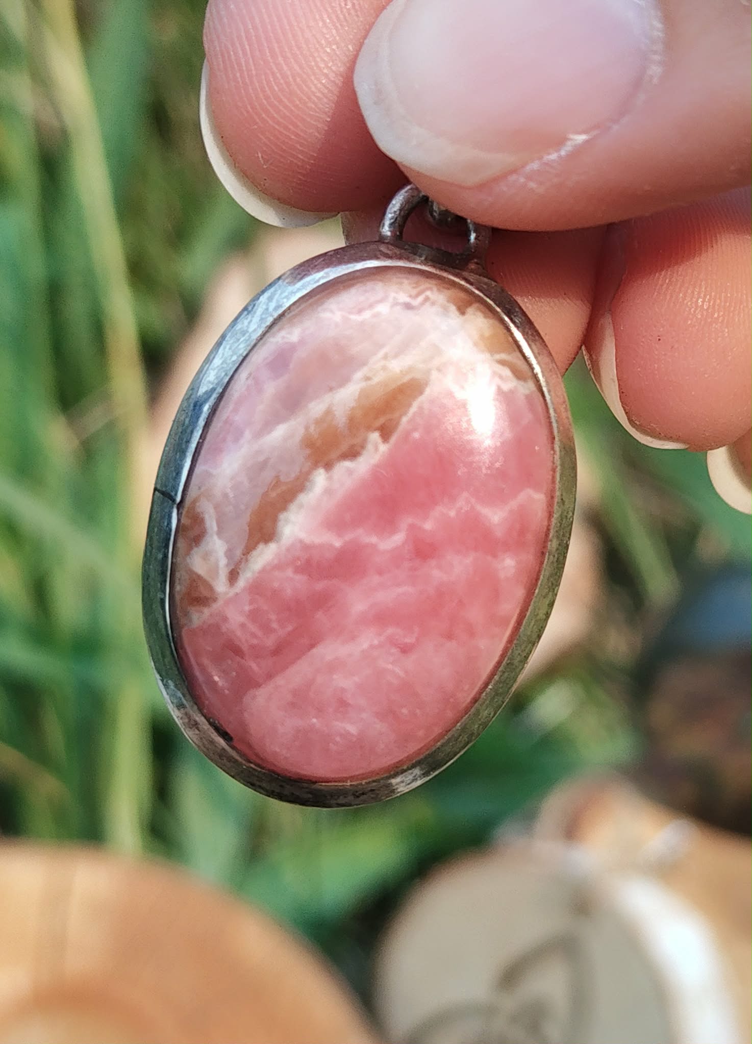 Pendentif rhodochrosite ovale rose marbré en argent 925 tenu entre les doigts devant un fond végétal, pierre naturelle associée à la délicatesse, au recentrage et à la paix émotionnelle.