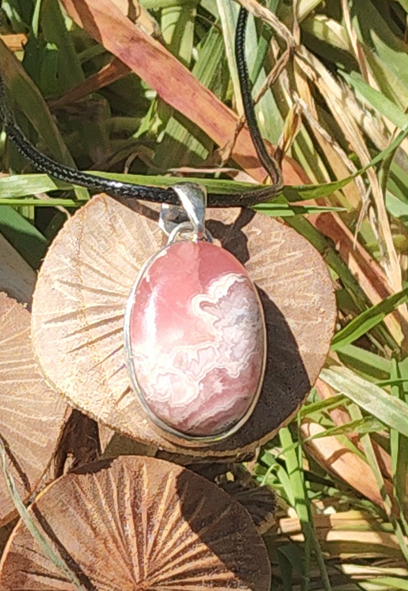 Pendentif rhodochrosite ovale rose marbré avec monture argent 925, posé sur un champignon et feuillage en lumière naturelle, pierre naturelle évoquant la tendresse, le soutien émotionnel et l’apaisement intérieur.