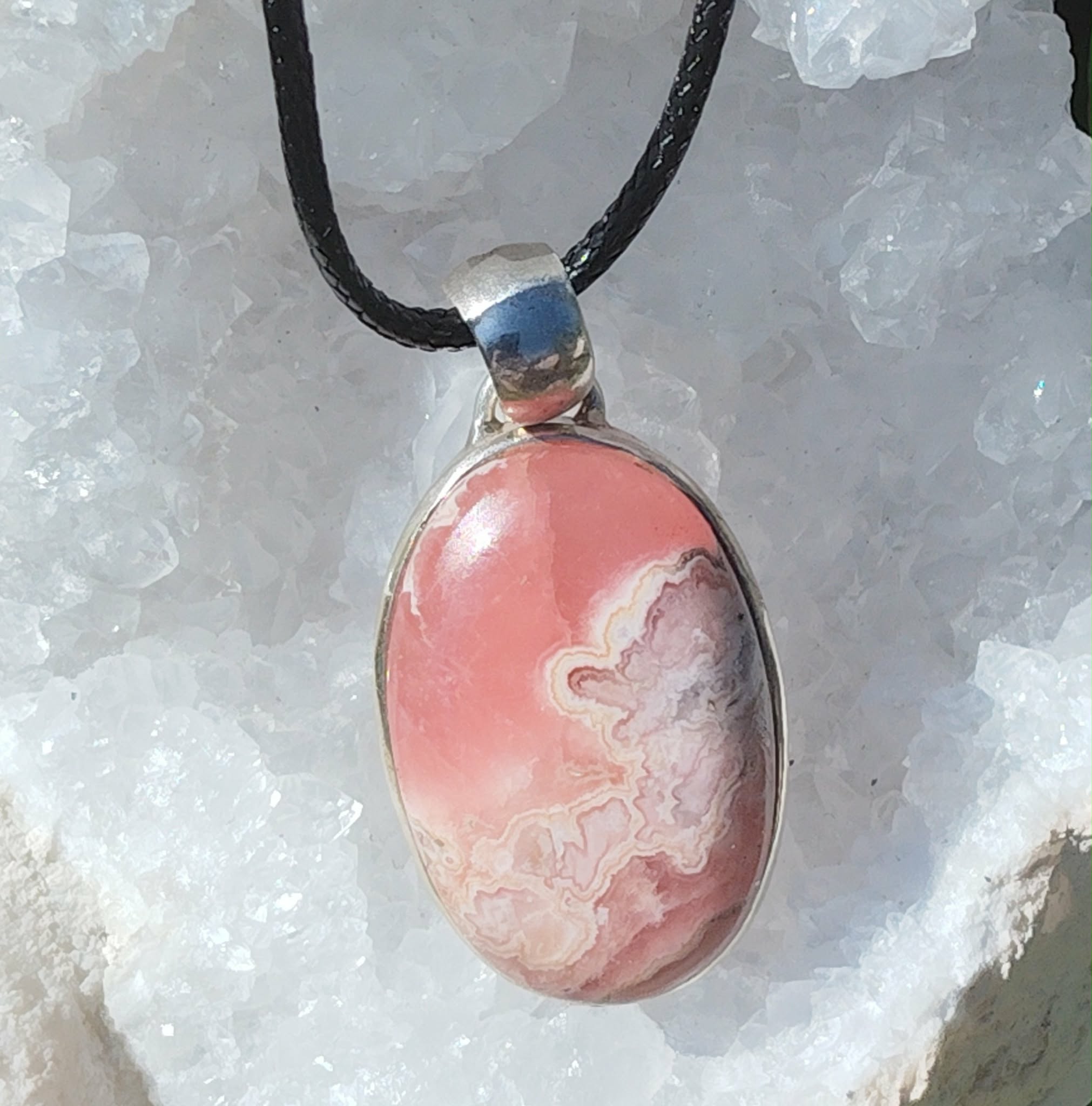 Pendentif rhodochrosite ovale rose marbré sur cordon noir, posé sur un amas de quartz, bijou en pierre naturelle associé à la tendresse, au réconfort et à l’équilibre émotionnel.