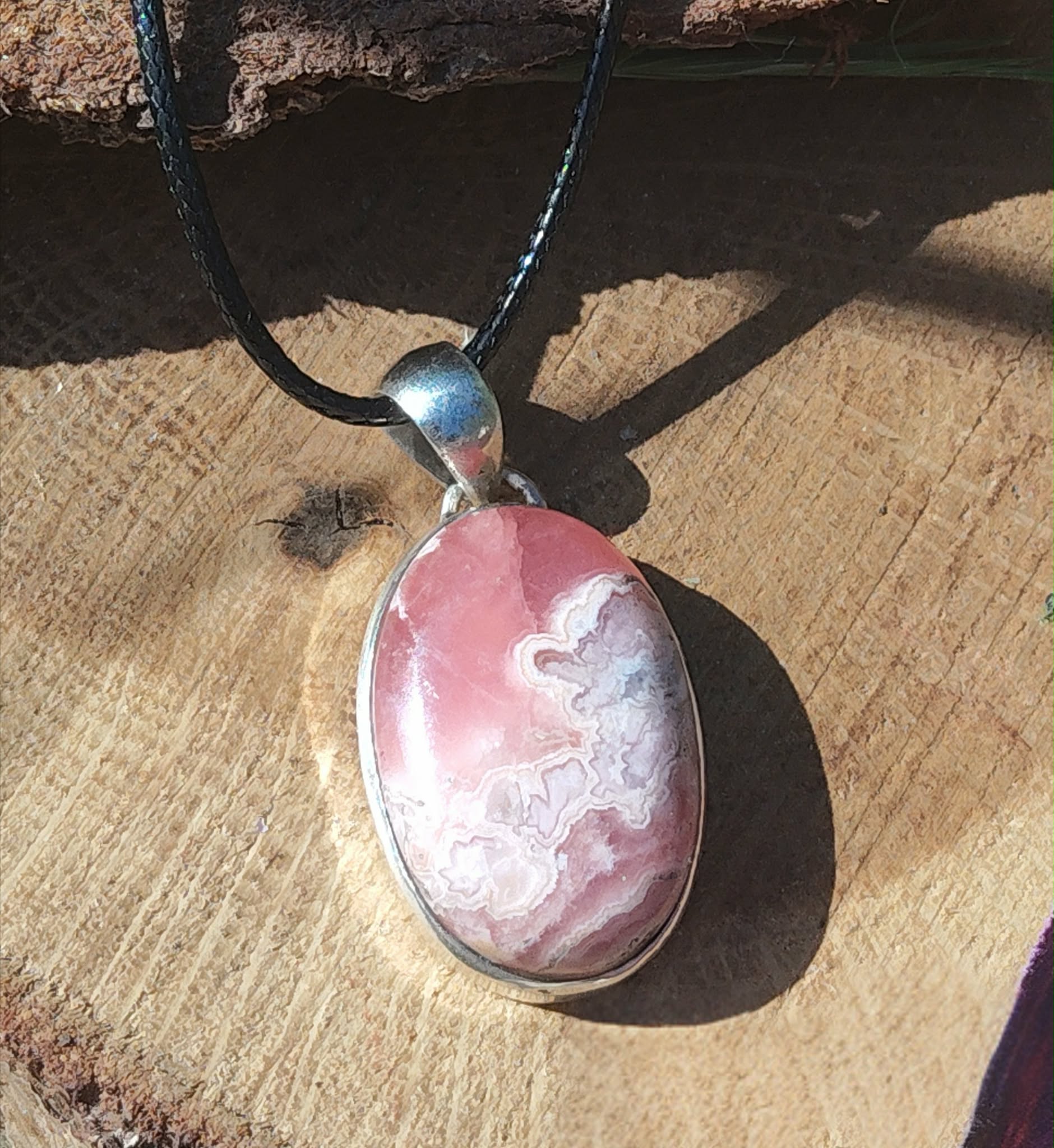 Pendentif rhodochrosite ovale rose marbré en argent 925 sur cordon noir, posé sur bois en lumière naturelle, bijou en pierre naturelle évoquant le calme affectif, la bienveillance et l’harmonie intérieure.