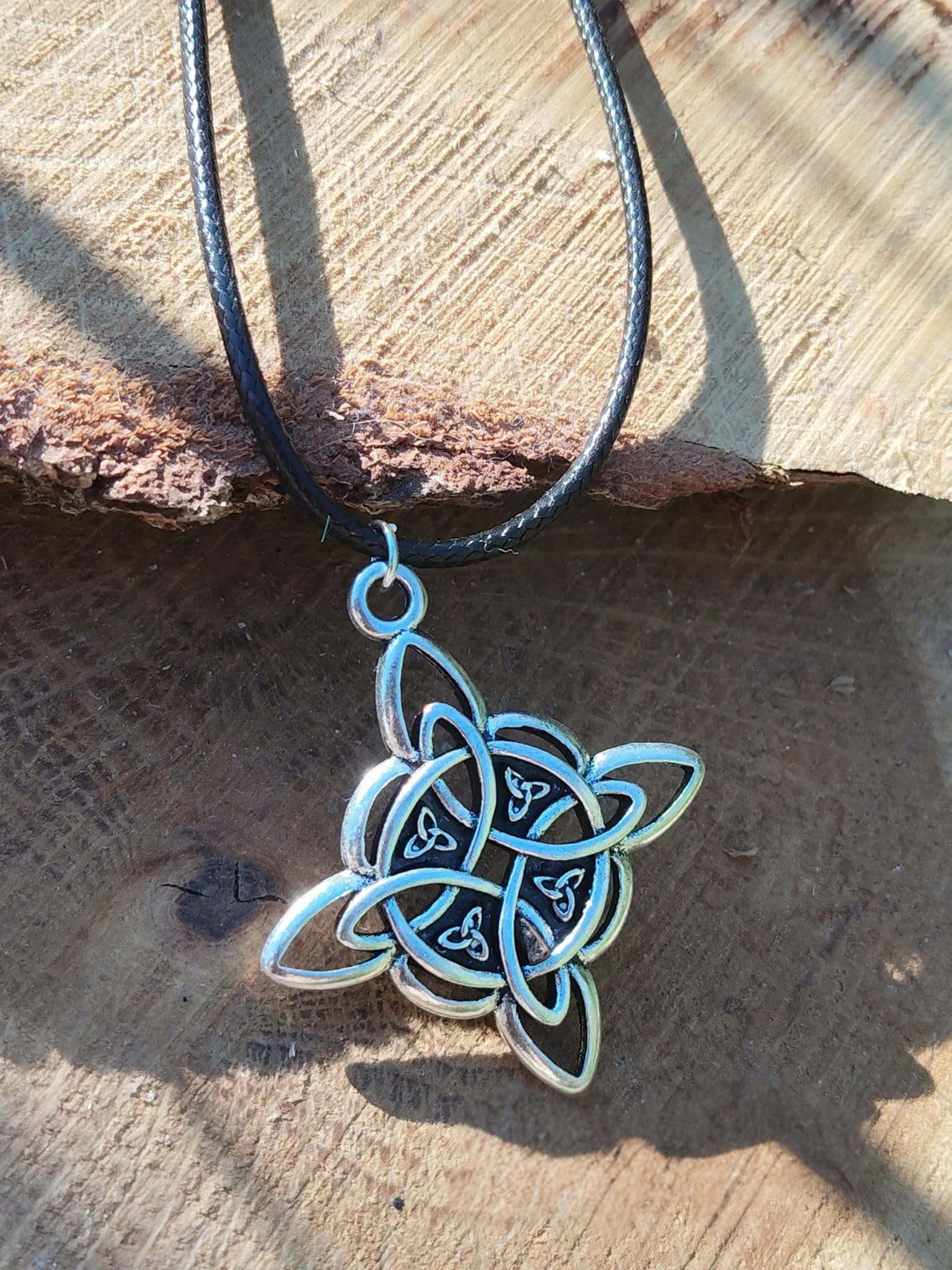 Collier sur cordon noir avec pendentif nœud de sorcière argenté, décor bois et écorce sombre en lumière naturelle, lien protecteur et style ésotérique