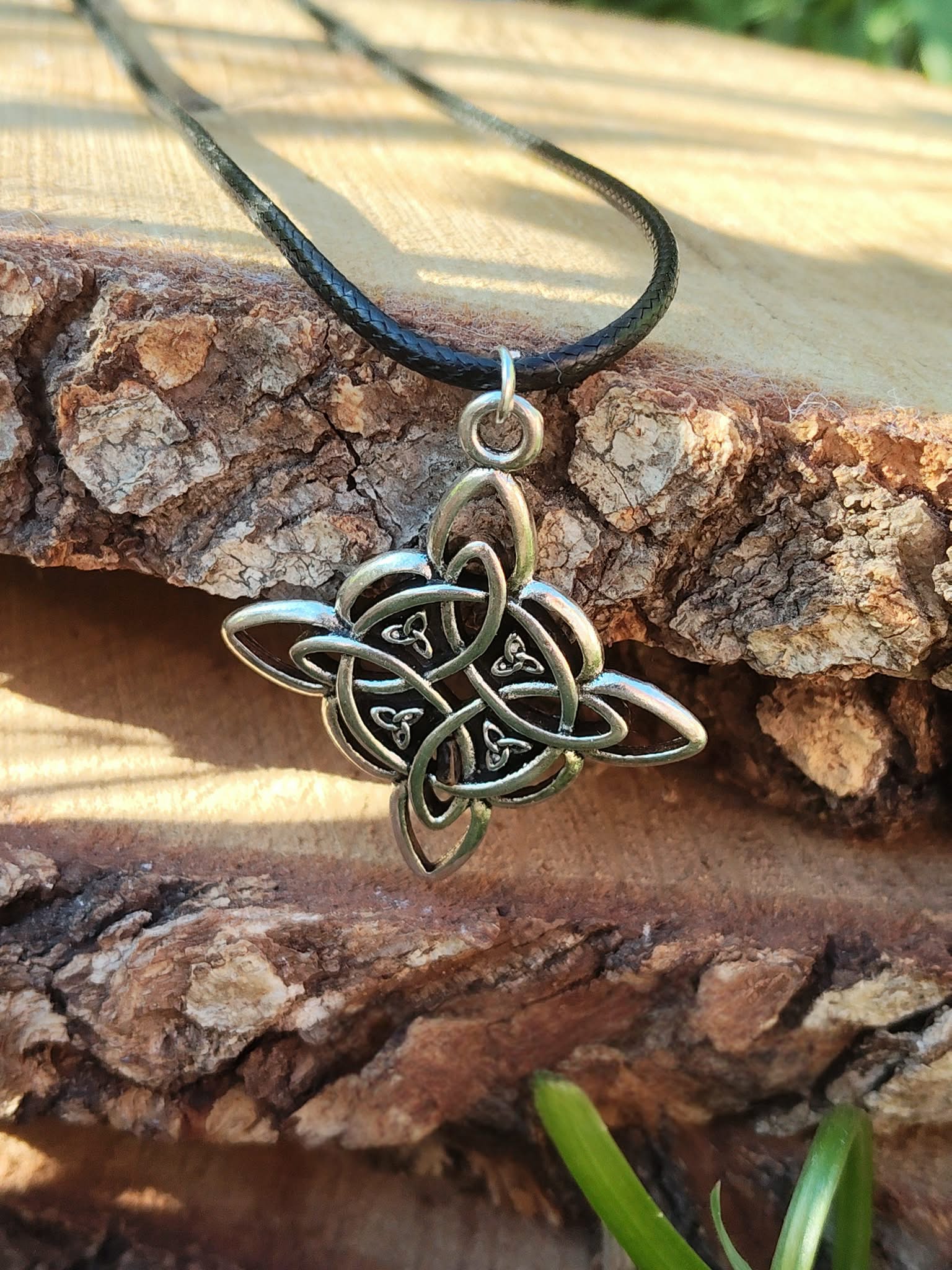 Collier nœud de sorcière argenté sur cordon noir, posé sur écorce et mousse avec feuillage, ambiance forêt et énergie protectrice