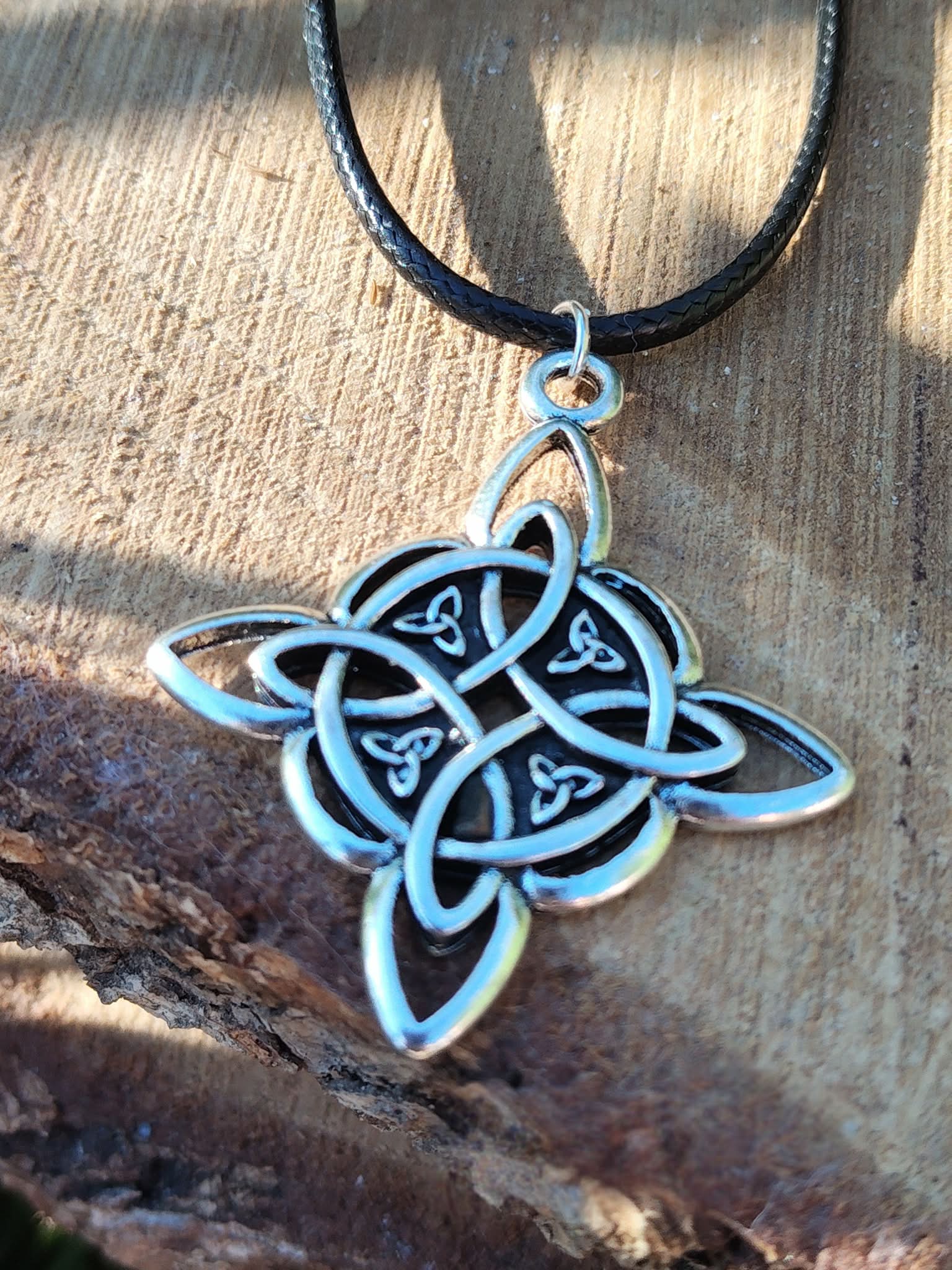 Gros plan sur pendentif nœud de sorcière argenté avec cordon noir, décor pierre claire et bois sombre, contraste minéral et protection symbolique