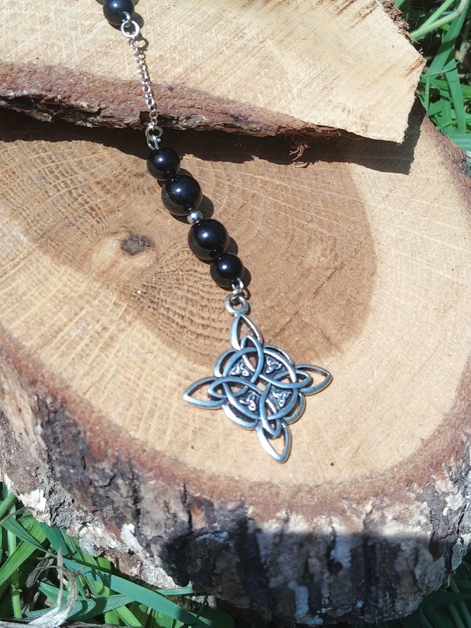 Bijou de rétroviseur « nœud de sorcière » : perles de tourmaline noire sur cordon, pendentif nœud celtique argenté, suspendu devant une tranche de bois et feuillage vert. Création Magic Gaïa, pour protection, ancrage et apaisement.