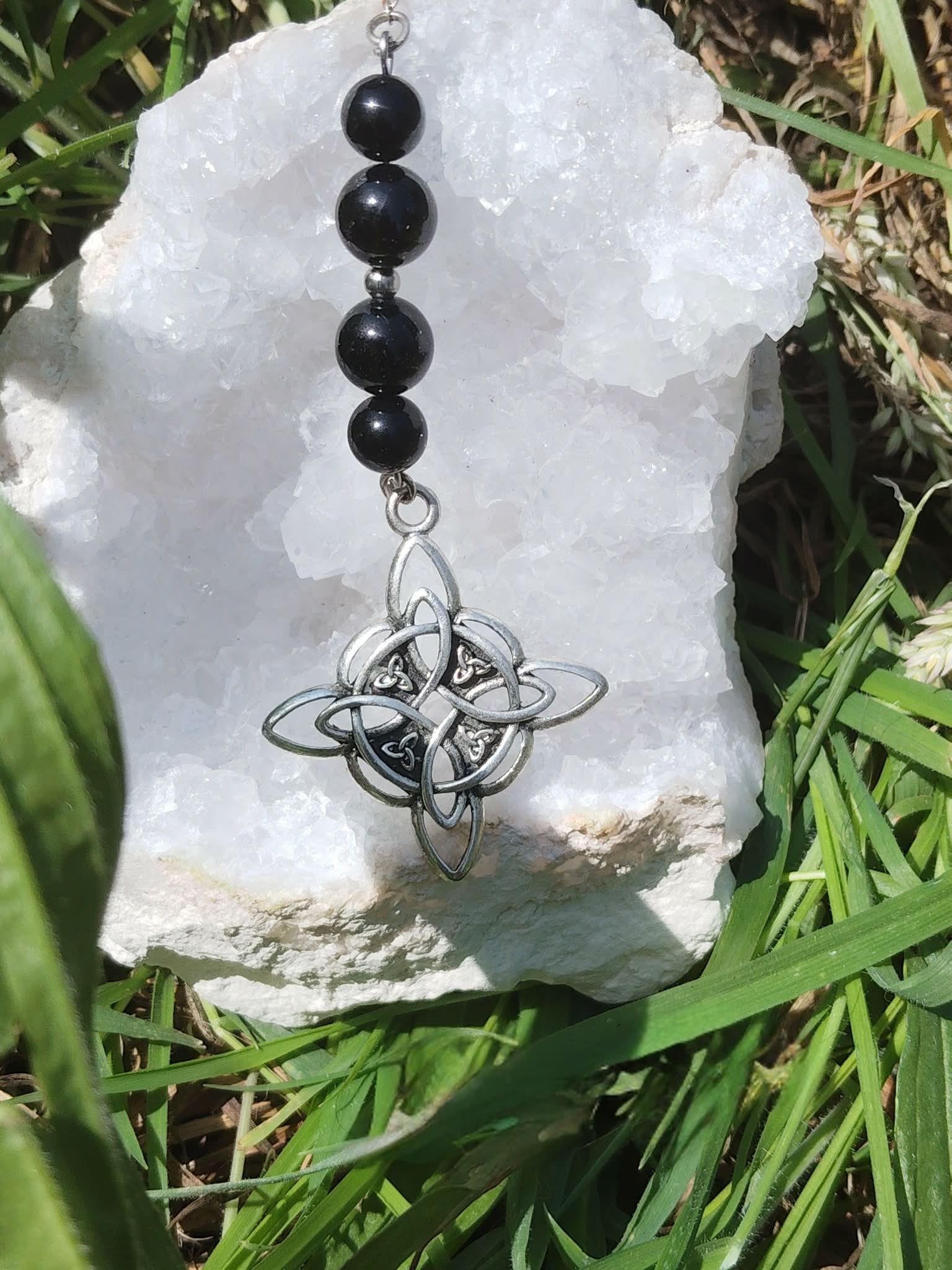 Bijou de rétroviseur « nœud de sorcière » : perles noires sur cordon, pendentif nœud celtique argenté, posé sur un amas de quartz blanc au milieu de feuillages verts. Création Magic Gaïa, pour protection, ancrage et apaisement.