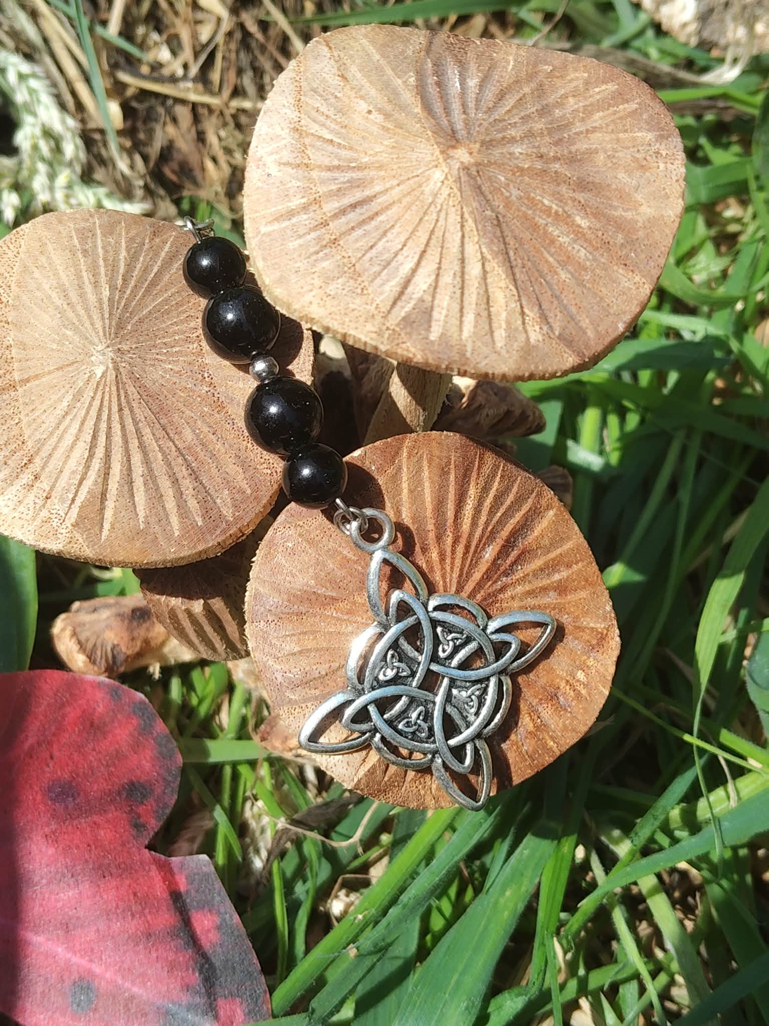 Bijou de rétroviseur nœud celte : perles de tourmaline noire et pendentif triquetra argenté, posé parmi champignons beige et feuillage vert. Création Magic Gaïa, pour protection, ancrage et sérénité.