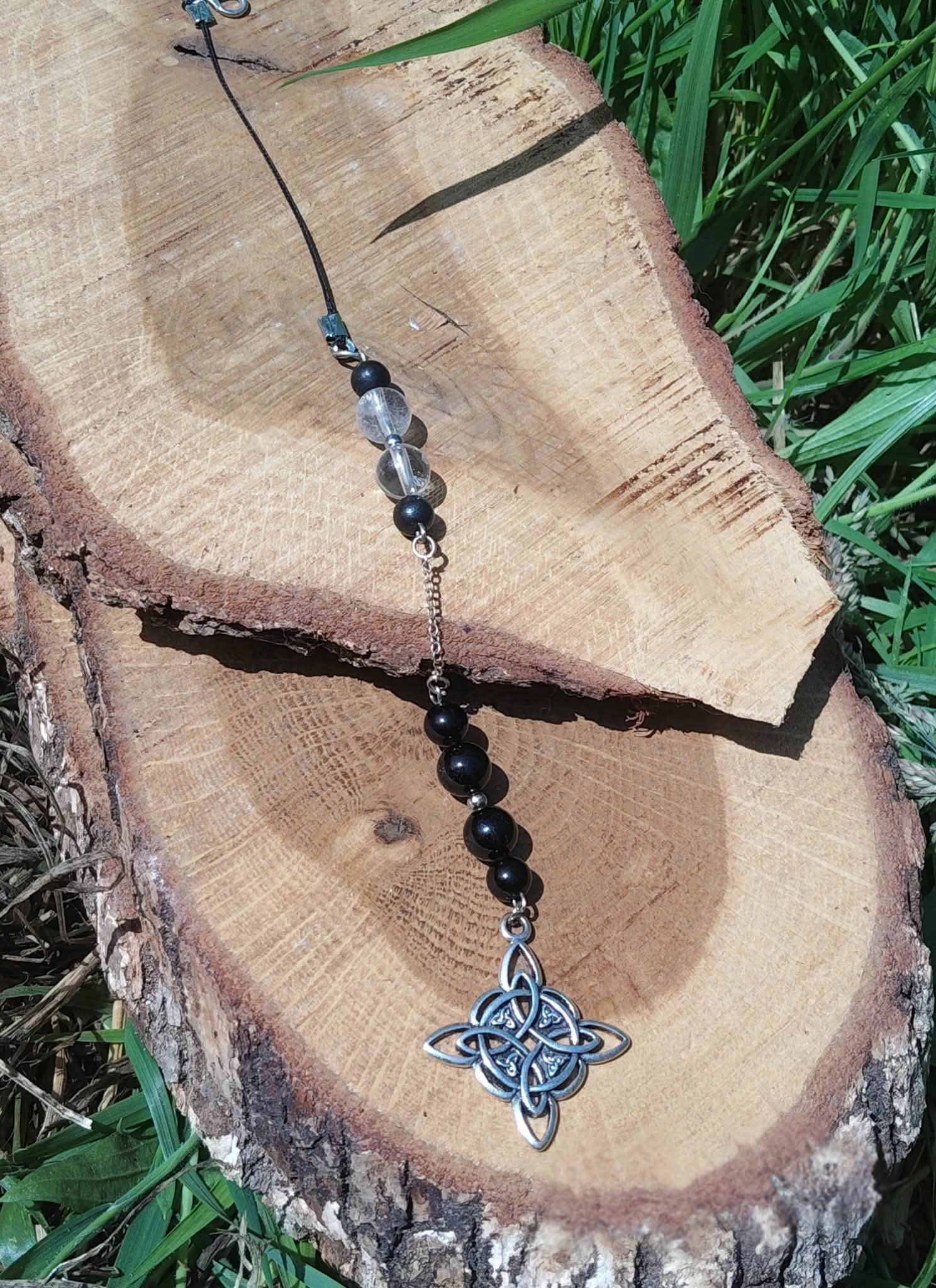 Bijou de rétroviseur « nœud de sorcière » : perles noires sur cordon bleu, pendentif nœud celtique argenté/bleuté, posé sur tranche de bois avec feuillage vert. Création Magic Gaïa, pour protection, ancrage et paix intérieure.