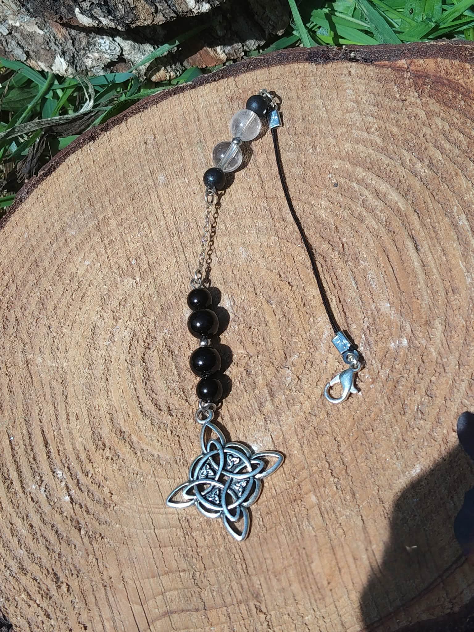 Bijou de rétroviseur « nœud de sorcière » : perles de tourmaline noire et petites perles claires sur cordon, pendentif nœud celtique argenté, posé sur rondin de bois avec feuillage vert. Création Magic Gaïa, pour protection, ancrage et apaisement.