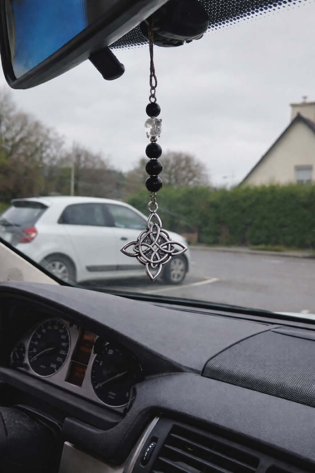 Bijou de rétroviseur « nœud de sorcière » : perles de tourmaline noire sur cordon, pendentif nœud celtique argenté au miroir. Photo dans la voiture, route en arrière-plan. Création Magic Gaïa, pour protection et ancrage.