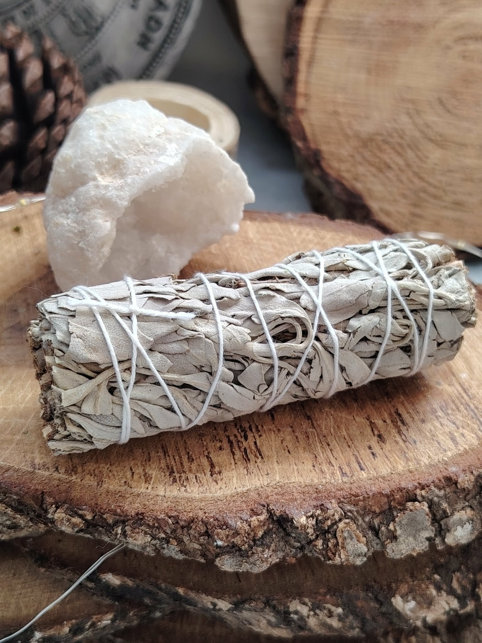 Bâton de sauge blanche à brûler (fumigation/smudge), fagot ficelé, posé sur rondin de bois avec amas de quartz et tranches de bois. Idéal pour rituel, méditation, purification énergétique, nettoyage des lieux, protection et apaisement