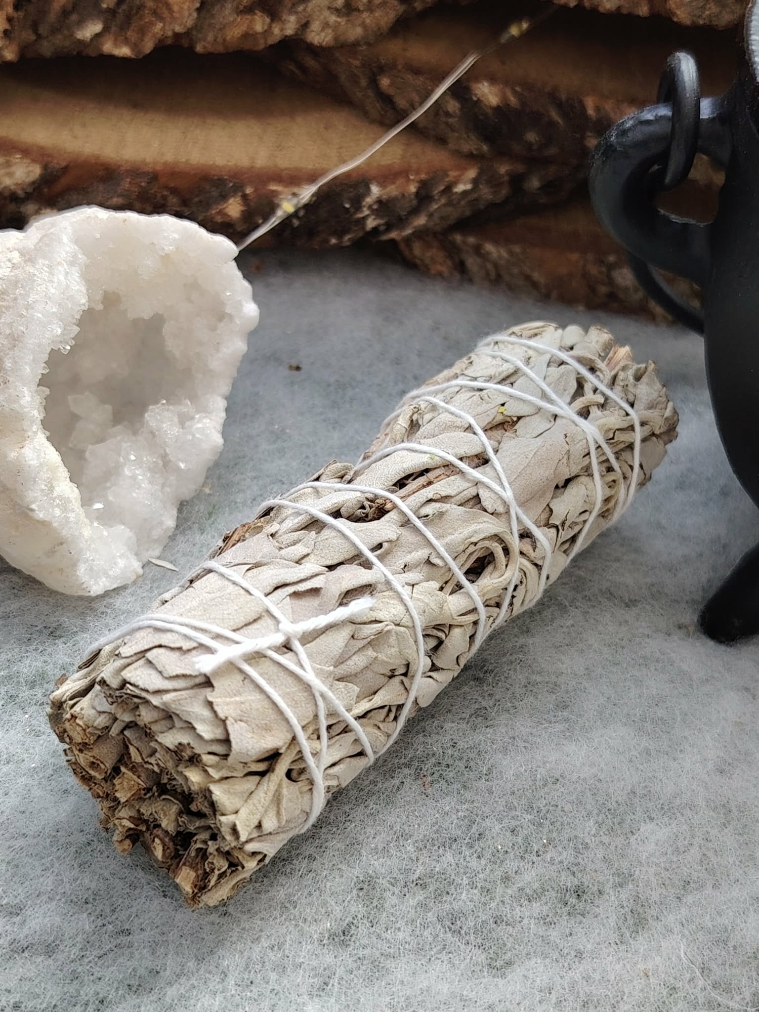 Bâton de sauge blanche à brûler (fumigation/smudge) ficelé, posé sur surface grise avec amas de quartz blanc. Accessoire ésotérique pour rituel, méditation, purification énergétique, nettoyage des lieux, protection, clarté et apaisement.