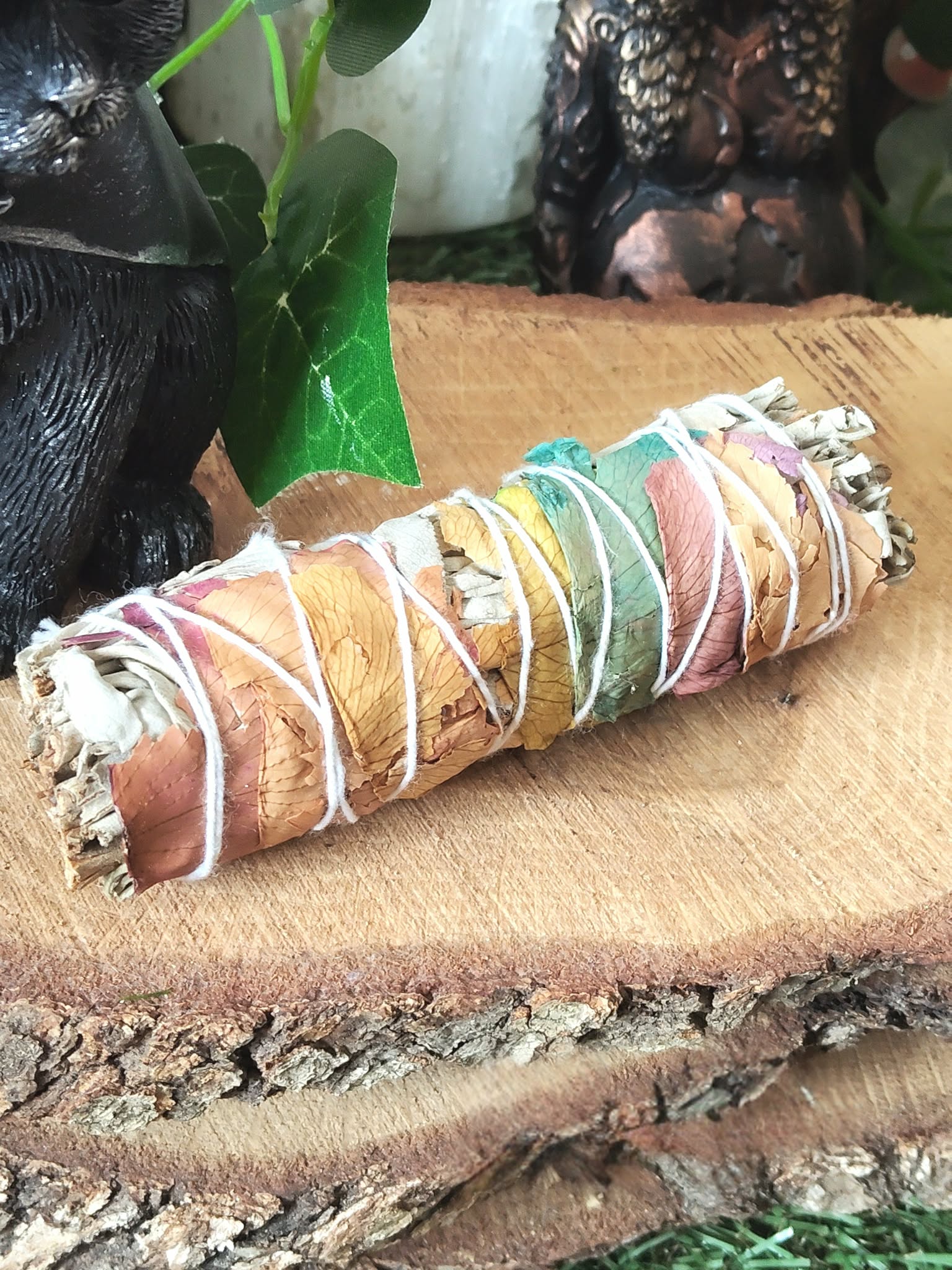 Bâton de sauge 7 chakras à brûler (fumigation/smudge), décoré de pétales multicolores, ficelé, posé sur rondin de bois avec chaudron noir et feuillage vert en arrière-plan. Purification énergétique, protection, clarté, apaisement.