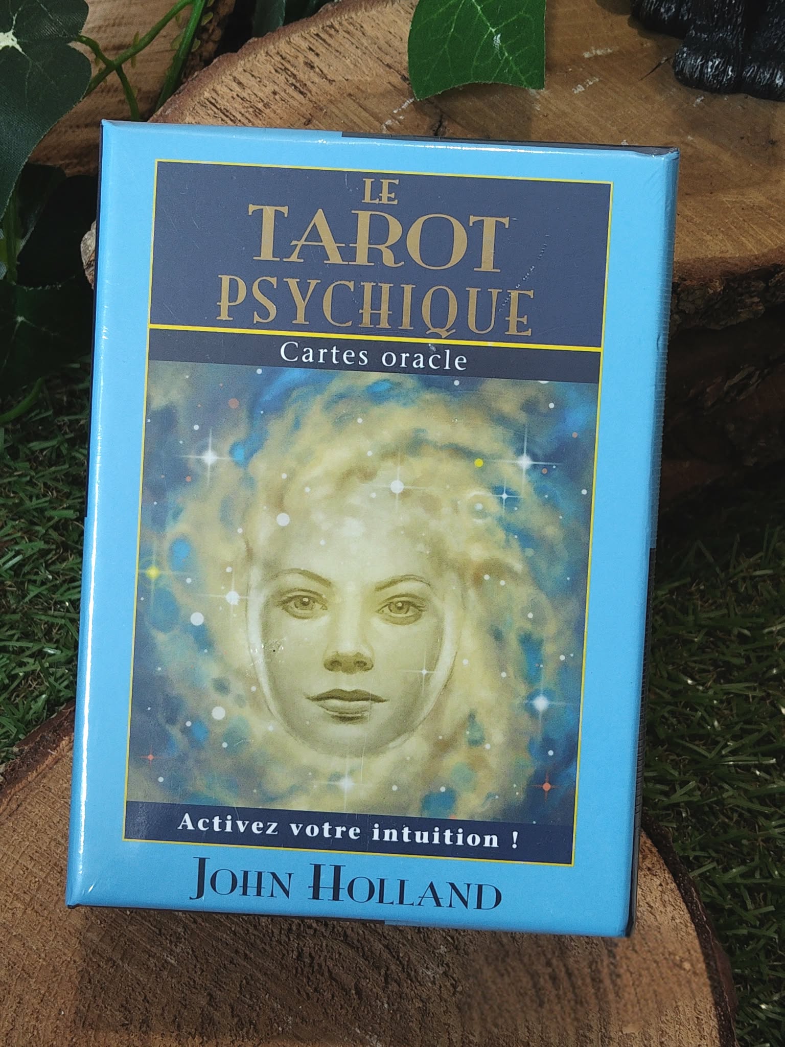 Boîte du coffret oracle « Le Tarot Psychique » de John Holland, étui bleu avec portrait, posée sur rondin de bois et feuillage. Cartes divinatoires pour tirage, guidance, intuition, méditation, développement personnel et clarté.