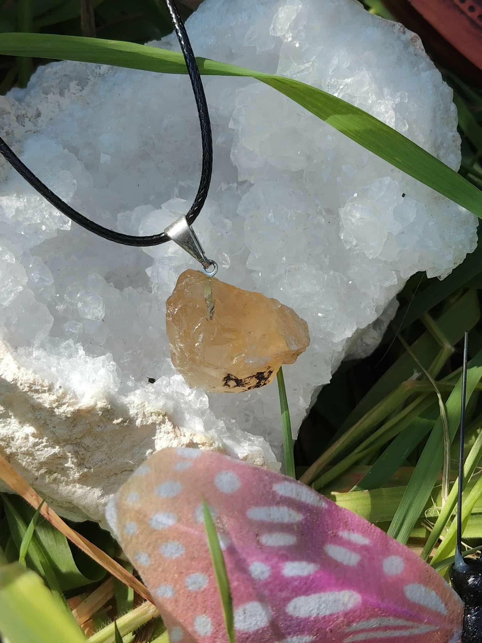 Pendentif citrine brute sur cordon noir, photo sur amas de quartz blanc avec brin d’herbe et carte rose à pois, pierre naturelle associée au dynamisme, à la lucidité et à l’énergie positive.