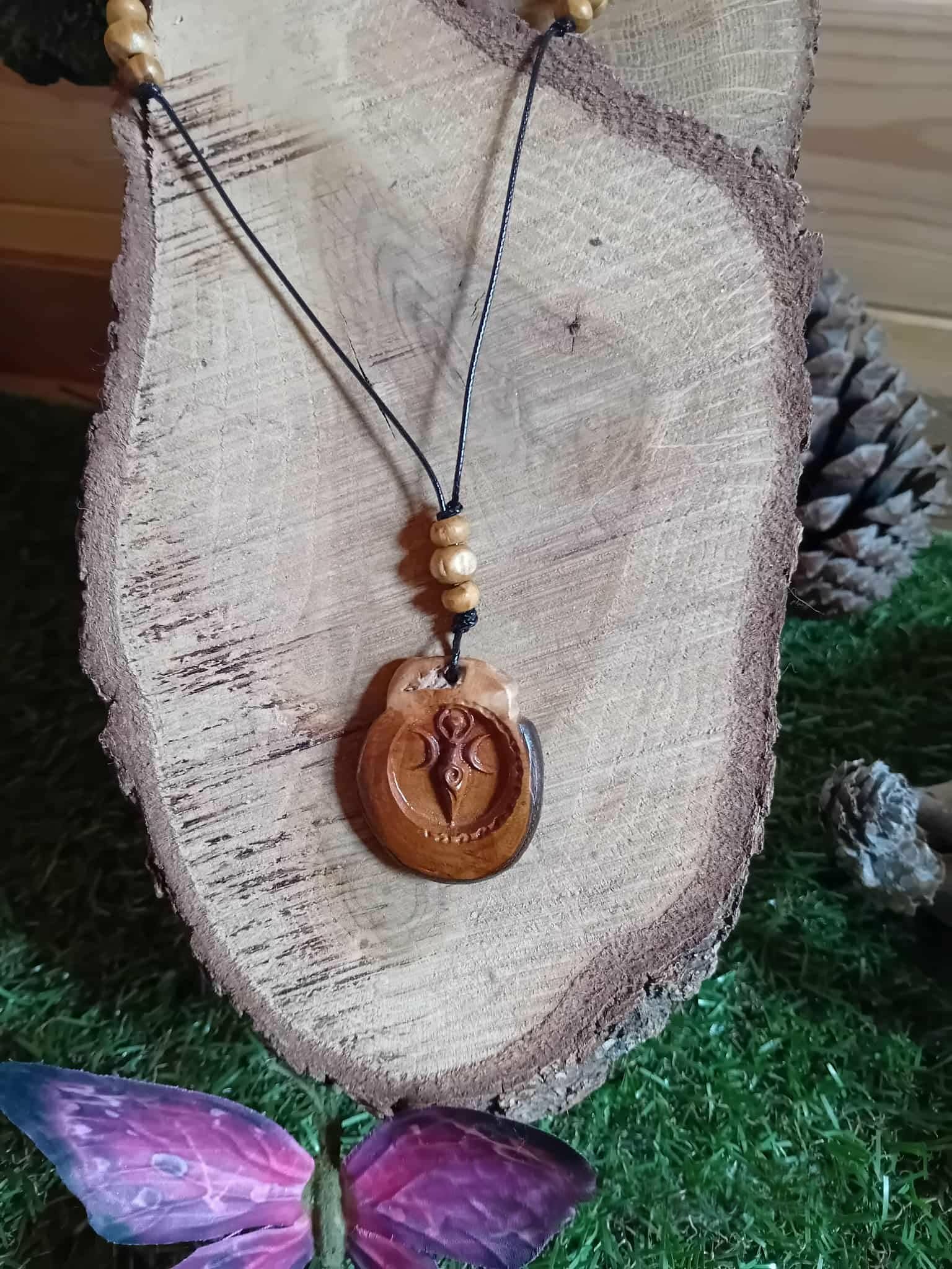 Pendentif bois Gaïa rond gravé suspendu devant tranche de bois avec mousse verte, cordon noir et perles bois dans une ambiance forestière