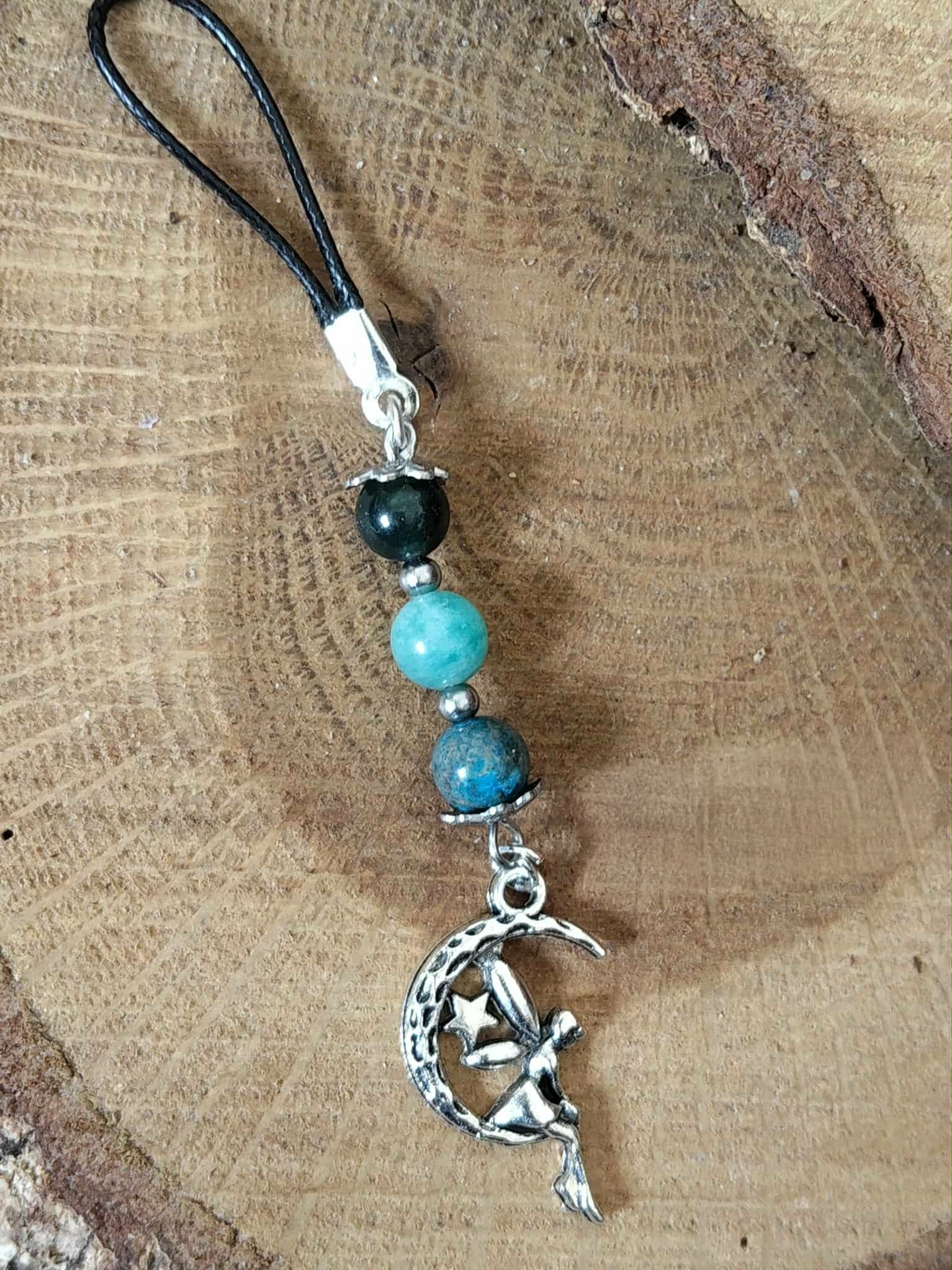 Bijou de téléphone création Magic Gaïa sur fond bois, cordon noir avec perles amazonite, agate mousse et aqua terra, breloque fée métal argenté, style bohème, apaisement, communication, ancrage, équilibre émotionnel.