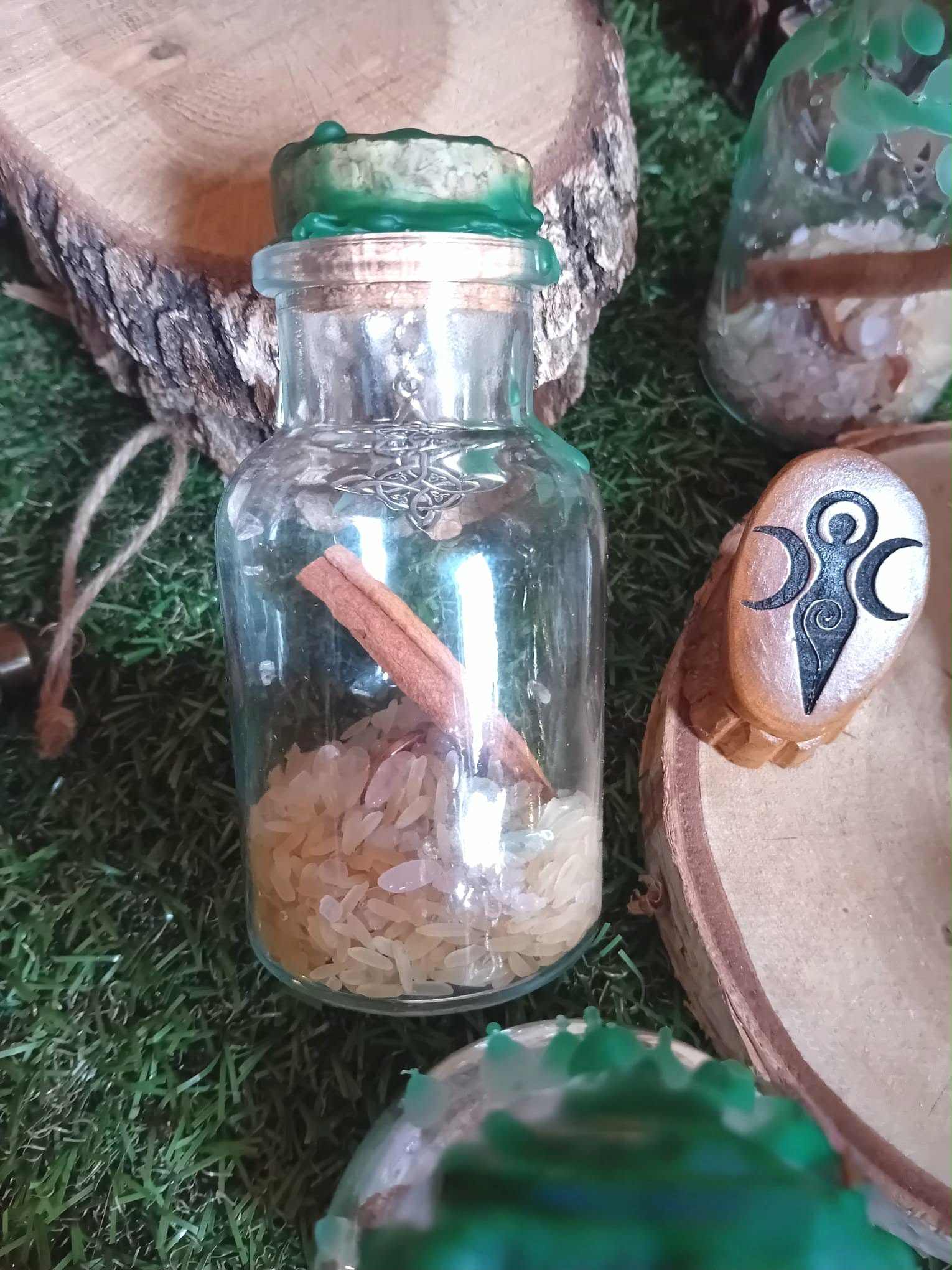 Fiole spell jar abondance Magic Gaïa, cire verte au bouchon, riz au fond et cannelle, décor nature mousse, tranche de bois en arrière-plan
