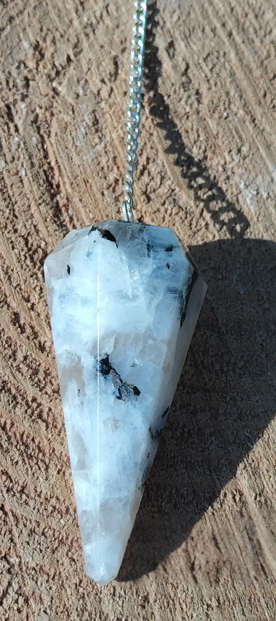 Pendule en péristérite (pierre de lune) facettée personnalisable sur demande, chaîne argentée, gros plan sur rondin de bois, décor naturel, radiesthésie/divination, intuition, apaisement, clarté, protection douce, équilibre émotionnel