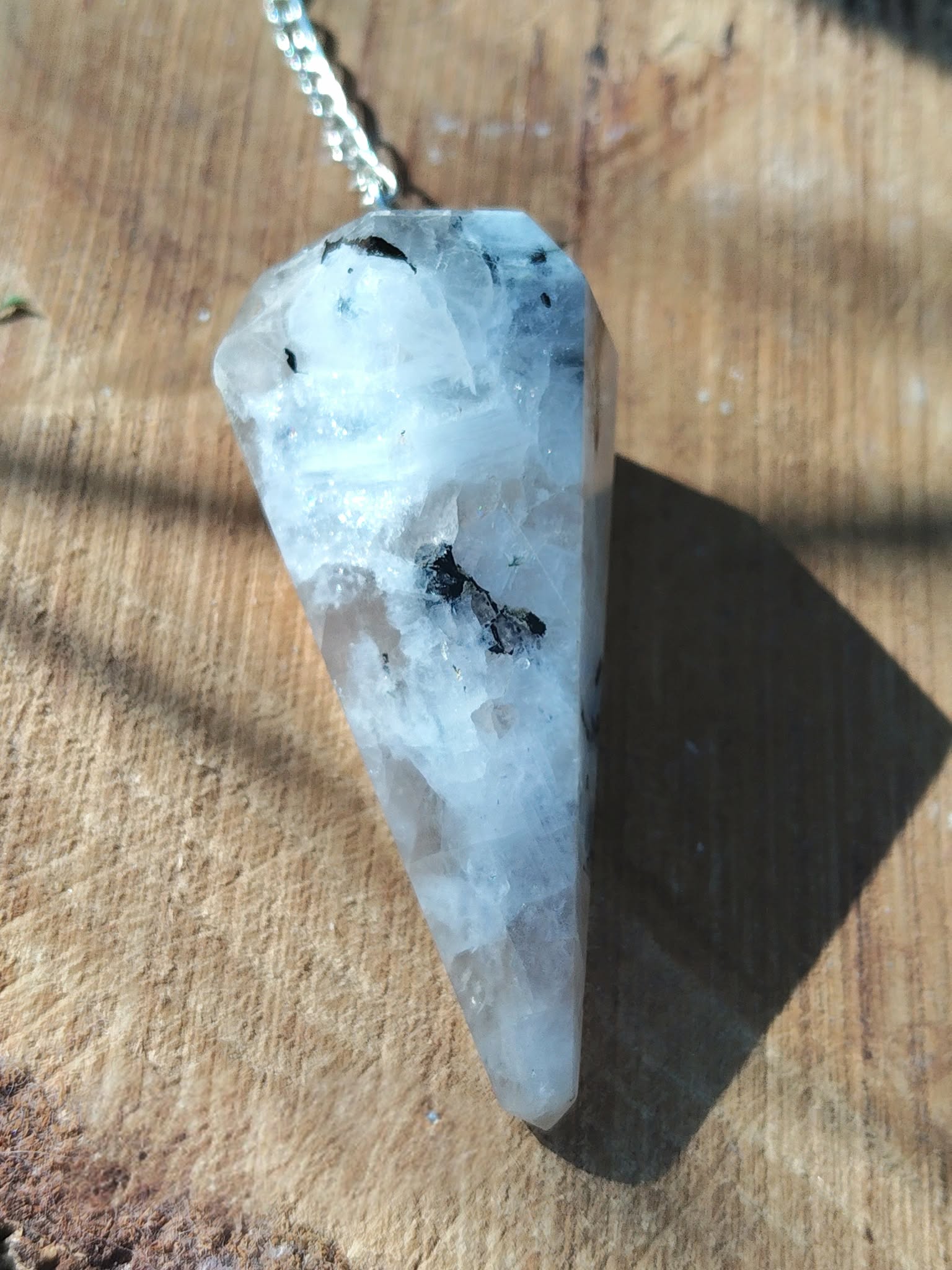 Pendule en péristérite (pierre de lune) facettée personnalisable sur demande, chaîne argentée, gros plan sur rondin de bois, décor naturel, radiesthésie/divination, intuition, apaisement, clarté, protection douce, équilibre émotionnel