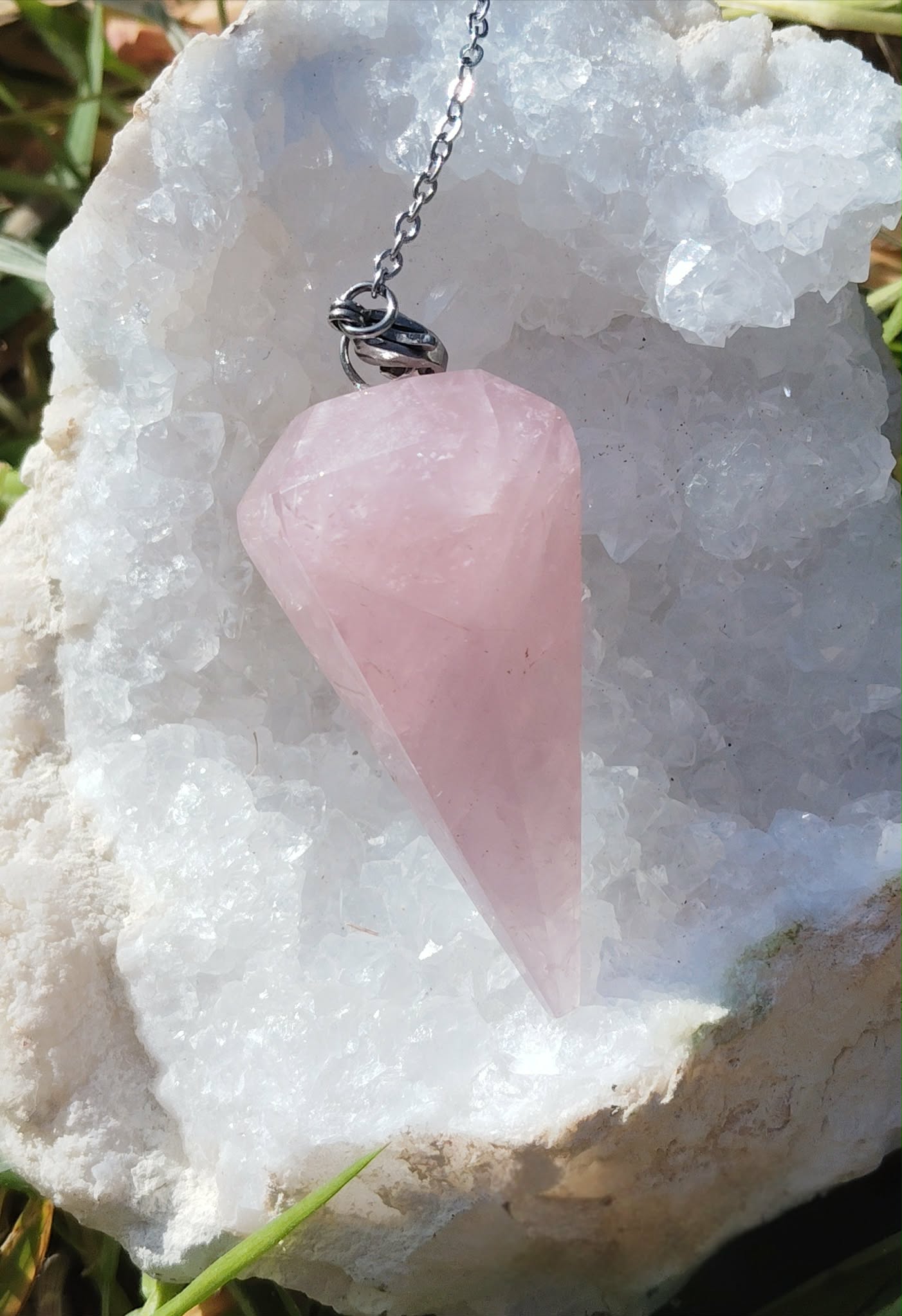 Pendule en quartz rose facetté personnalisable sur demande, chaîne argentée, posé sur amas de quartz blanc, décor naturel (mousse/herbes), radiesthésie/divination, douceur, apaisement, amour de soi, réconfort, harmonie émotionnelle