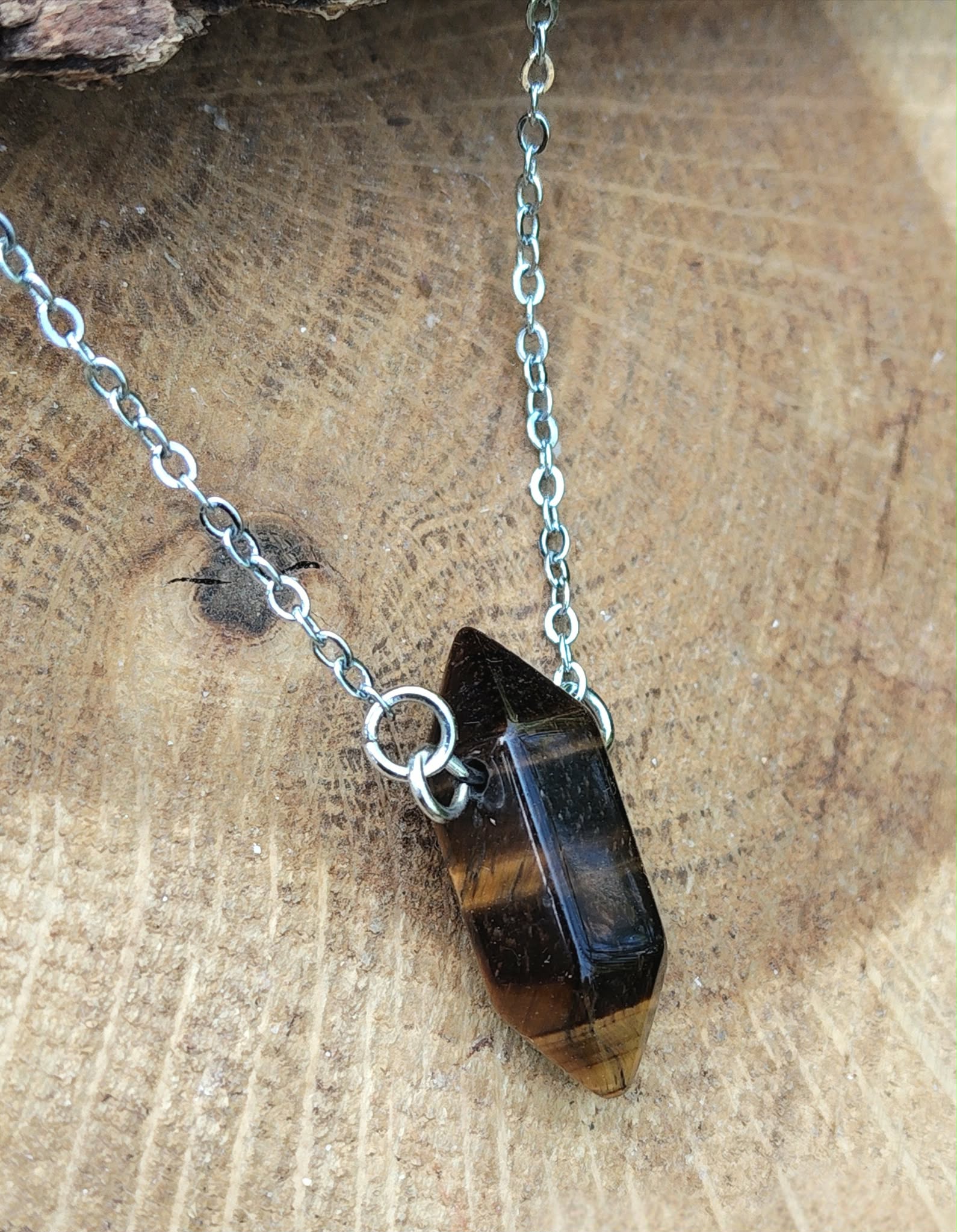Pendentif pointe œil de tigre sur chaîne en acier inoxydable, posé sur fond bois brut, bijou en pierre naturelle évoquant la force calme, la volonté et l’équilibre émotionnel.