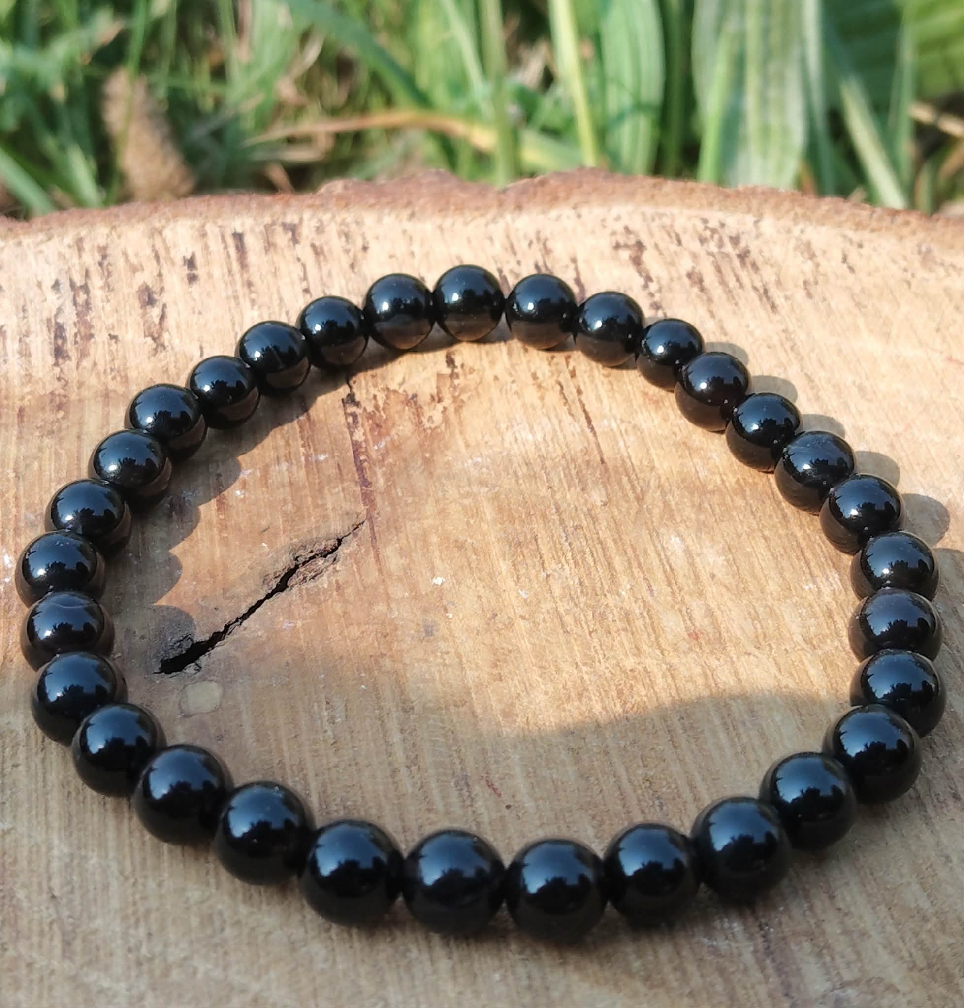 Bracelet Onyx noir en perles rondes sur élastique, posé sur tranche de bois avec herbes vertes, aplomb, constance et sang-froid.