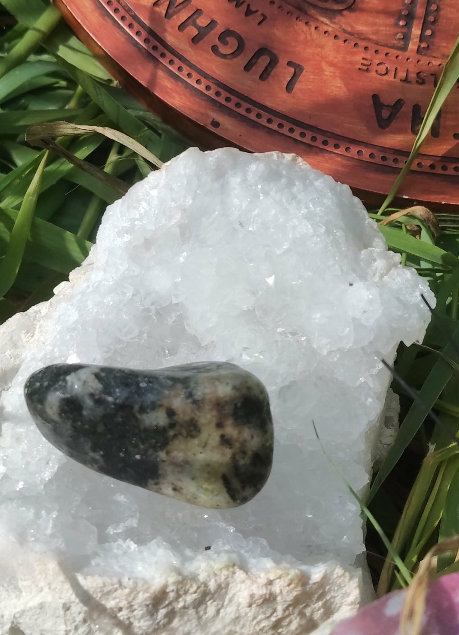 Pierre roulée de serpentine vert olive sur amas de quartz blanc, feuillage en fond; apaisement, ancrage, équilibre émotionnel.