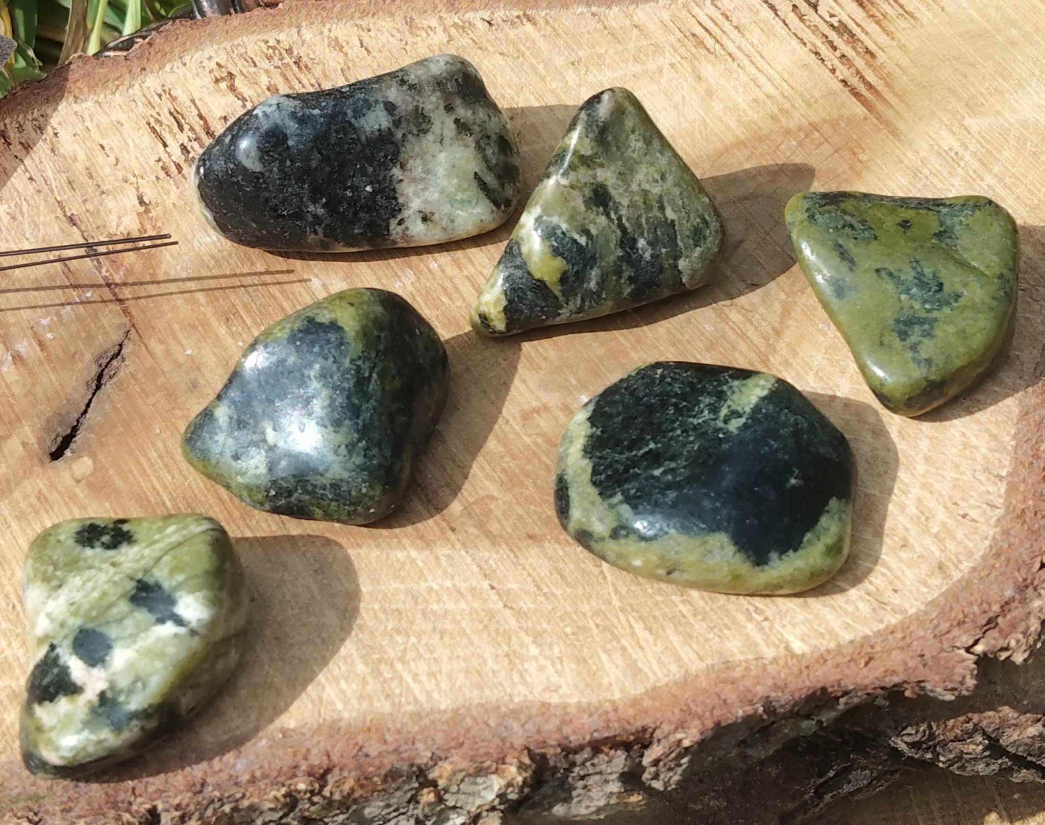Pierres roulées de serpentine vert olive, posées sur une tranche de bois clair en décor naturel, lumière douce et chaleureuse; pierre associée à l’apaisement, à l’ancrage et à l’équilibre émotionnel.