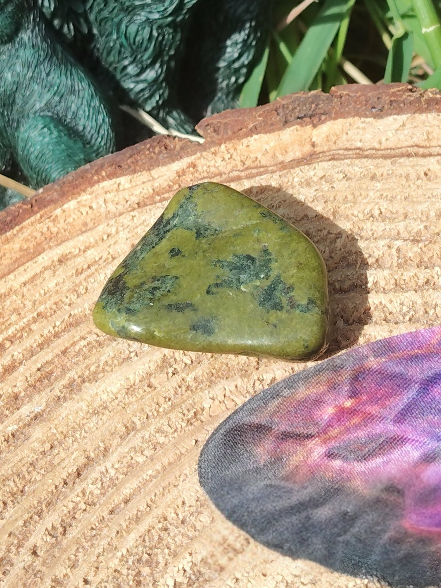 Pierre roulée de serpentine vert olive, sur tranche de bois avec plume violette et feuillage, lumière douce; associée à l’apaisement, à l’ancrage et à l’équilibre émotionnel.
