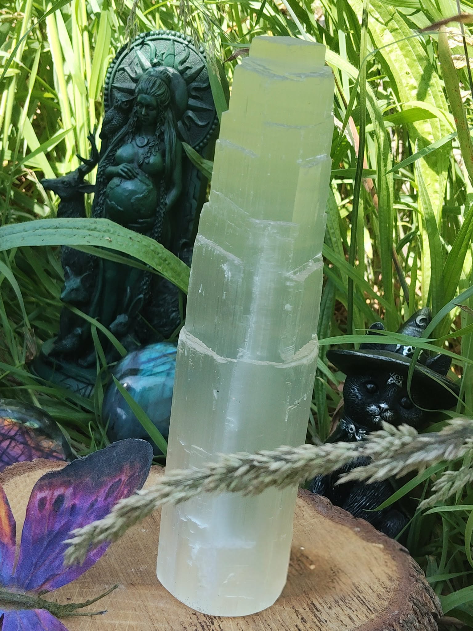 Tour en sélénite blanche posée dans l’herbe, fond nature, corde naturelle devant, plume multicolore et petit élément bleu à côté, sensation de purification douce.