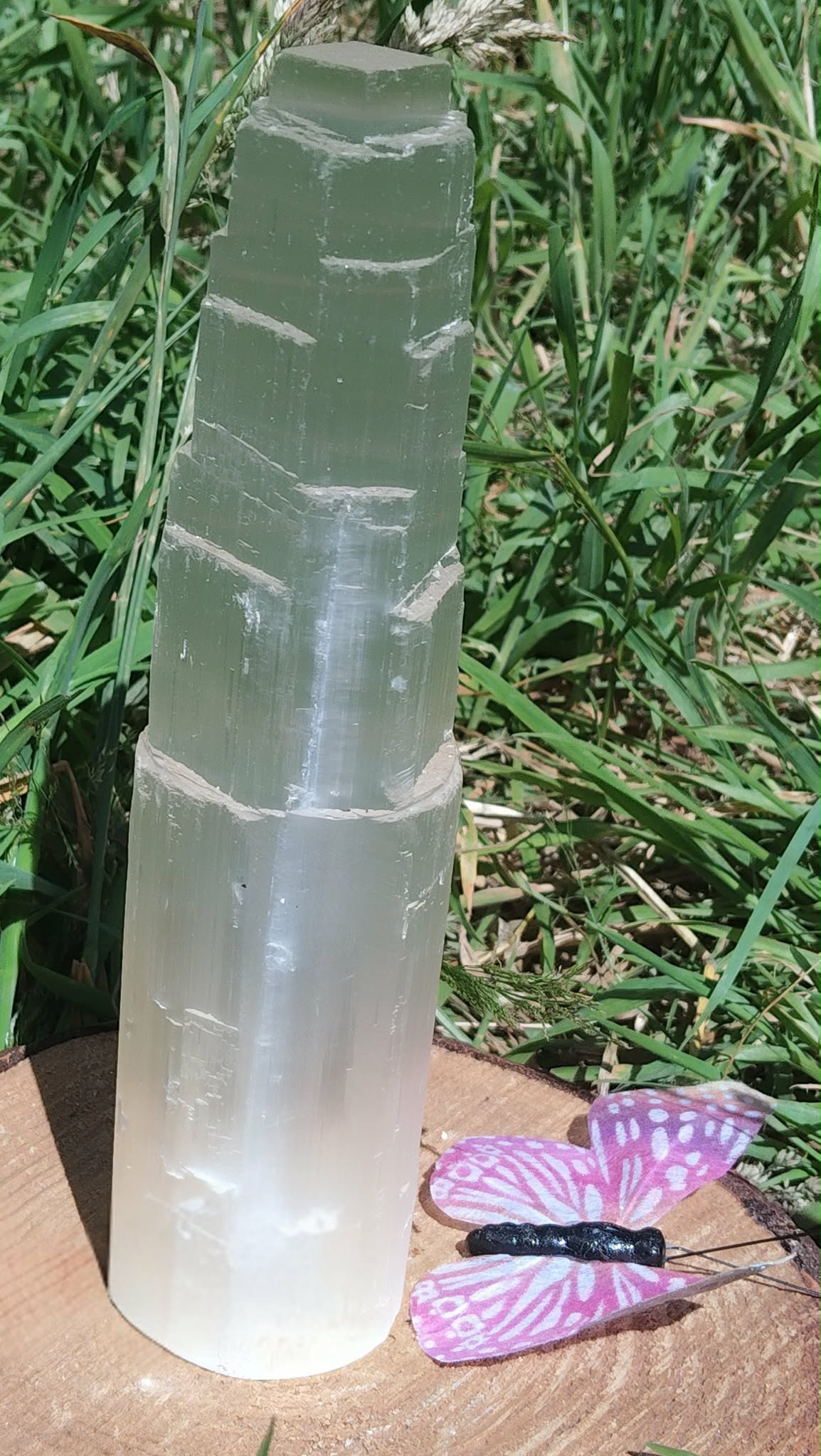 Tour en sélénite translucide photographiée en extérieur sur fond d’herbe, avec un papillon violet au bas de l’image, ambiance douce et apaisante.