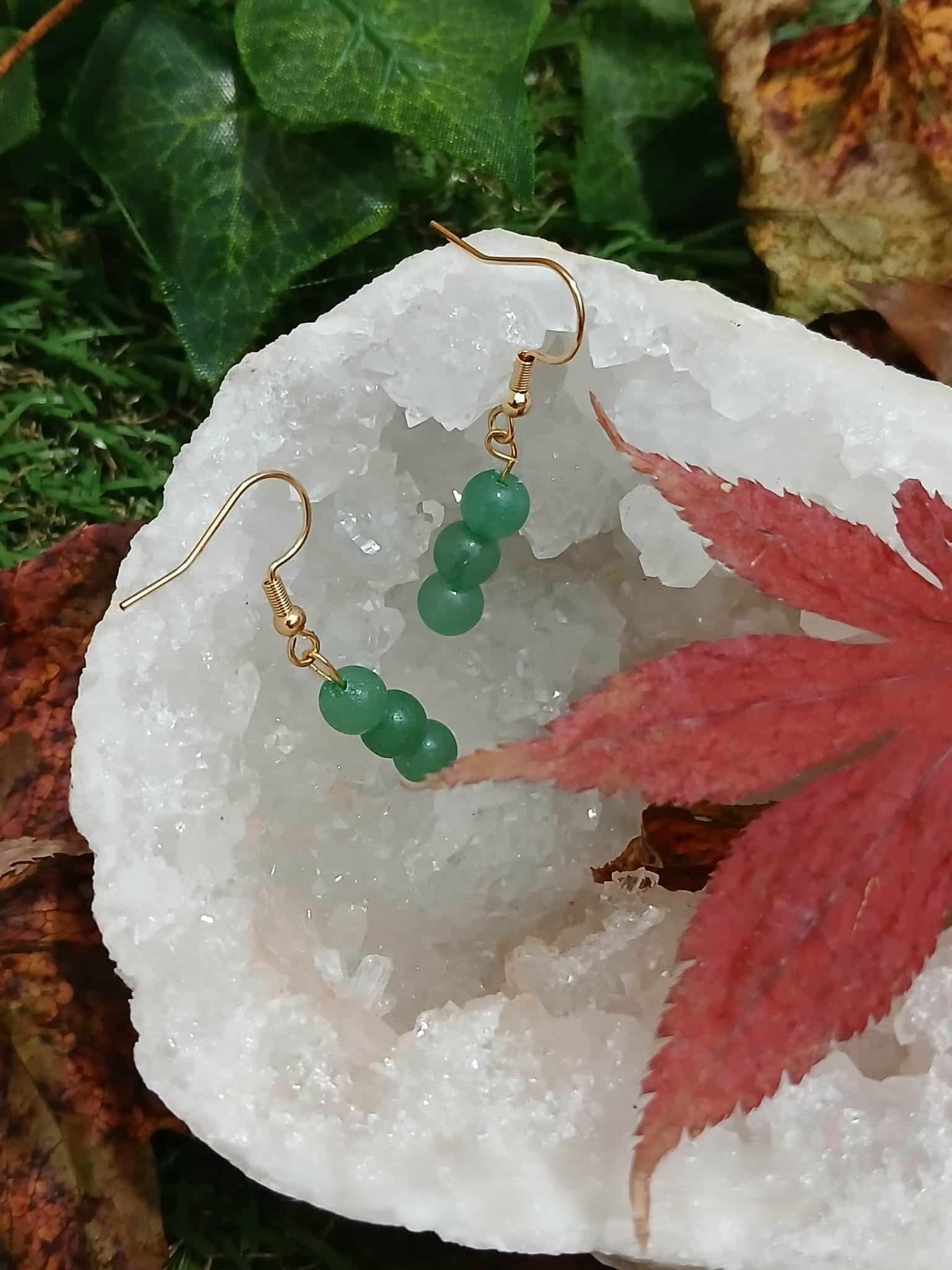 Création Magic Gaïa : boucles d’oreilles en aventurine verte sur crochets dorés, posées sur amas de quartz blanc avec feuille rouge, fond végétal, pour apaisement et harmonie.
