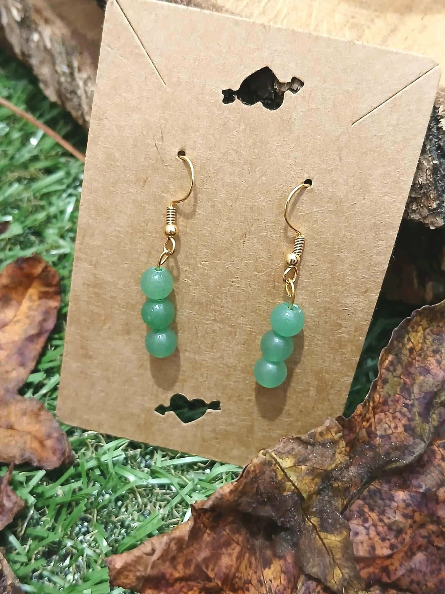 Boucles d’oreilles création Magic Gaïa en aventurine verte, posées sur amas blanc avec feuille rouge, fond végétal, pour une ambiance harmonieuse et sereine.