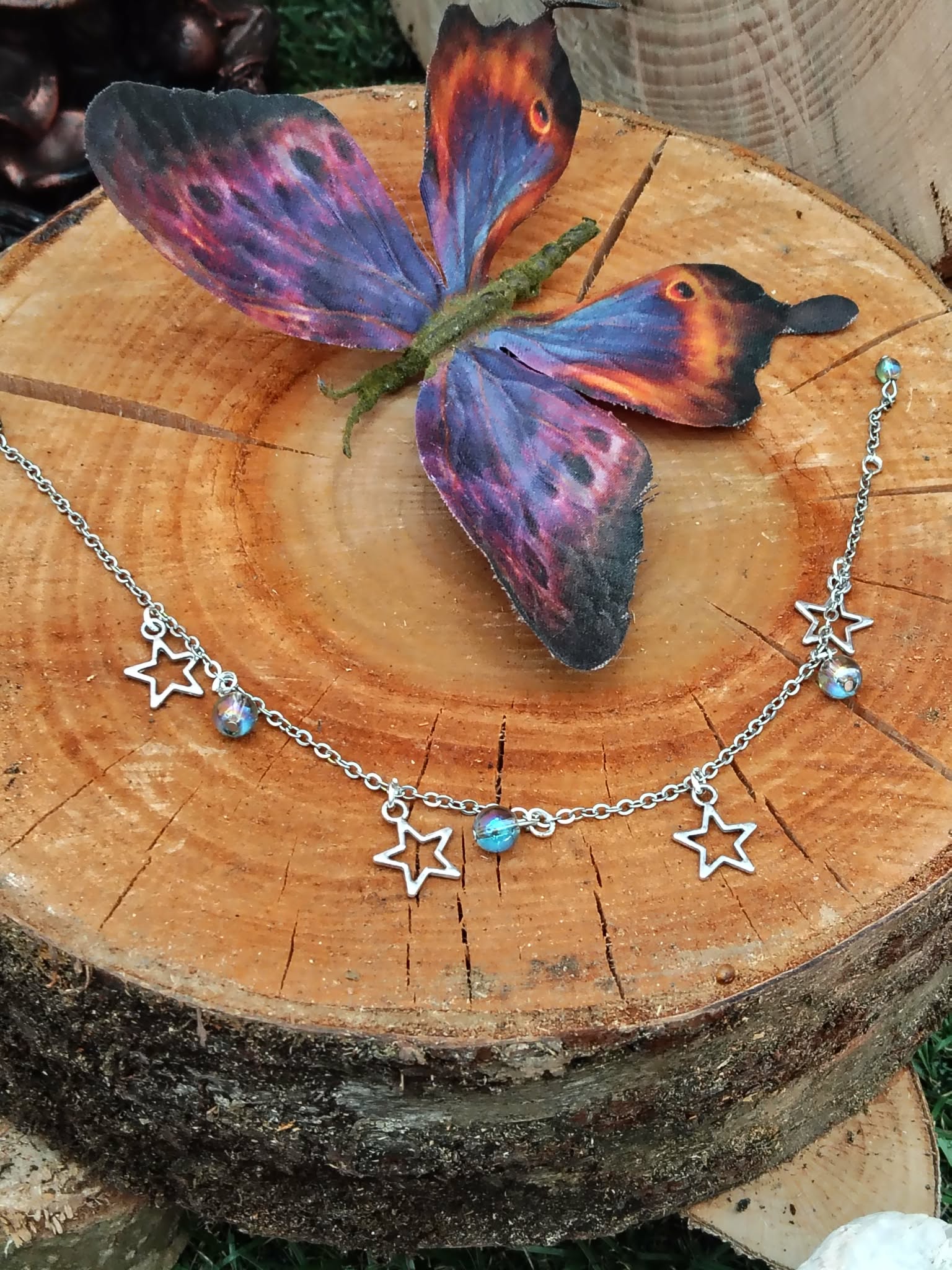 Chaîne de cheville création Magic Gaïa en acier inoxydable, étoiles et perles Aqua Aura, posée sur un rondin de bois avec un papillon coloré, pour une touche de fraîcheur et confiance.