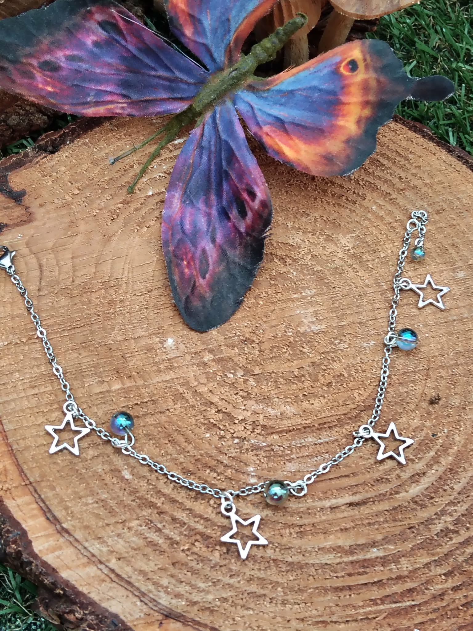 Chaîne de cheville création Magic Gaïa en acier inoxydable, étoiles et perles Aqua Aura, posée sur rondelle de bois avec papillon bleu-violet, pour légèreté et confiance.