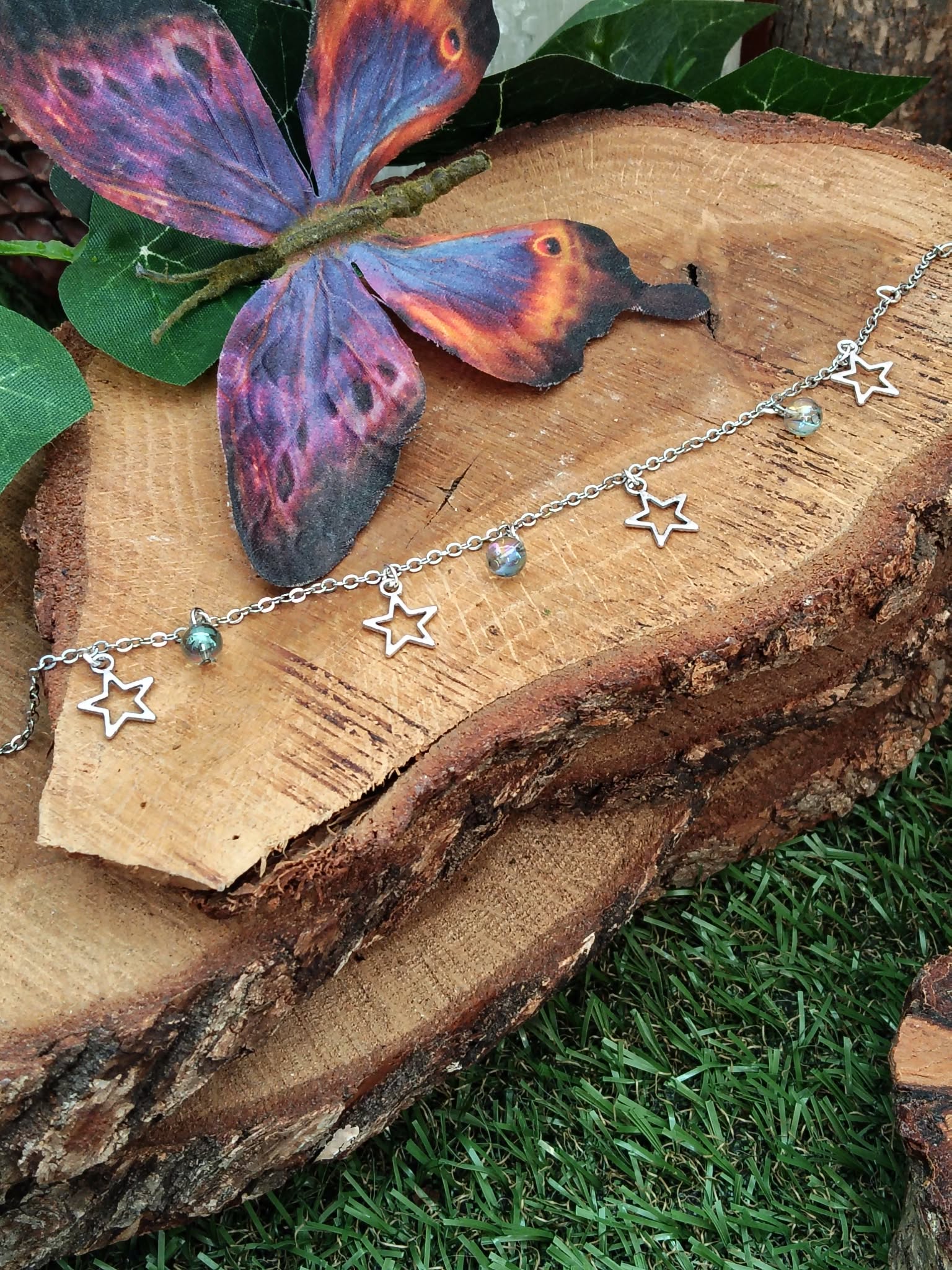 Chaîne de cheville création Magic Gaïa en acier inoxydable avec étoiles et perles Aqua Aura, posée sur rondin de bois avec papillon bleu-violet et mousse, pour une touche de légèreté.