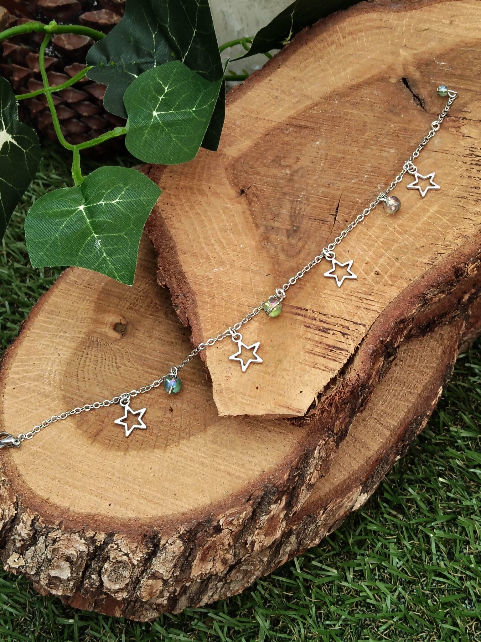 Chaîne de cheville création Magic Gaïa en acier inoxydable, étoiles et perles Aqua Aura, posée sur rondins de bois empilés avec mousse et feuilles de lierre, pour une énergie fraîche et assurance