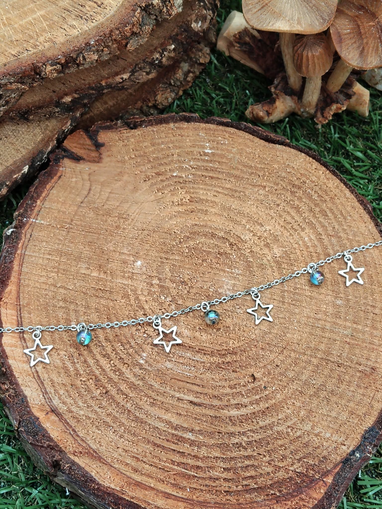 Chaîne de cheville création Magic Gaïa en acier inoxydable, étoiles et perles Aqua Aura, posée sur rondin de bois sur mousse avec petits champignons, pour une touche de fraîcheur et confiance.