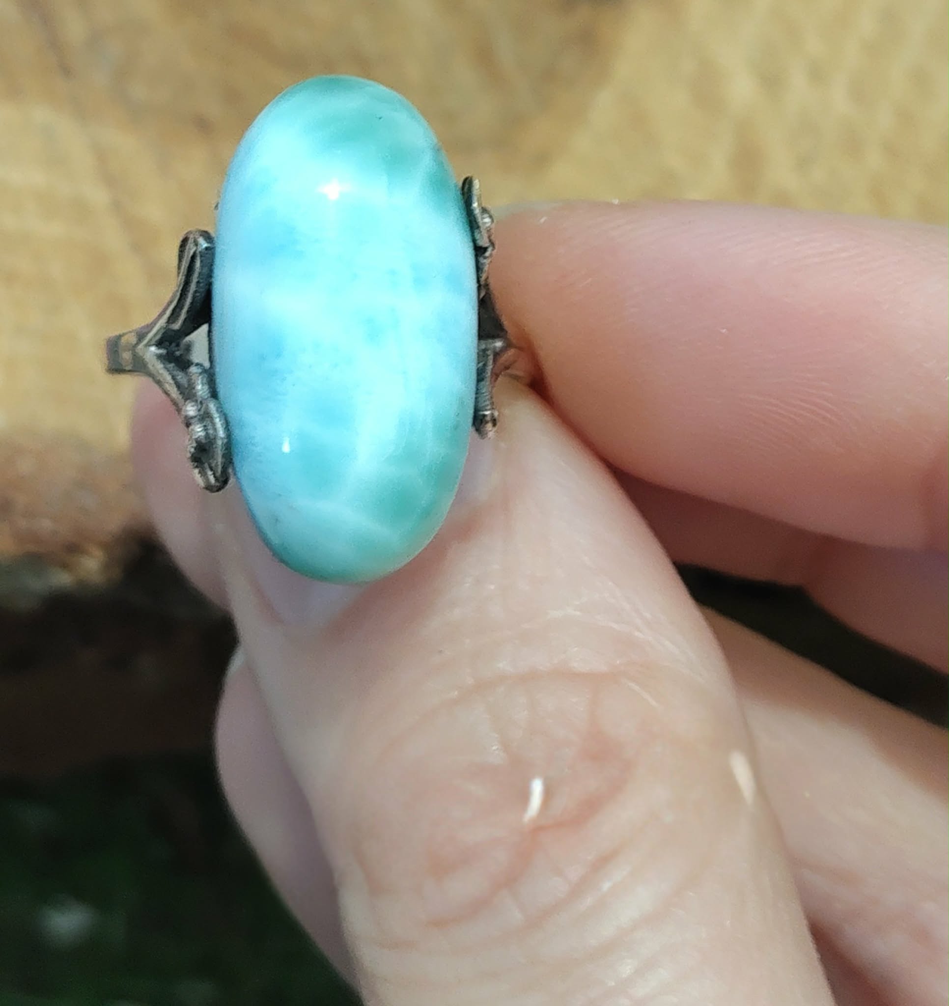 Bague larimar réglable en argent, tenue entre deux doigts devant un fond en bois doré, pour apaiser et encourager une expression sereine.