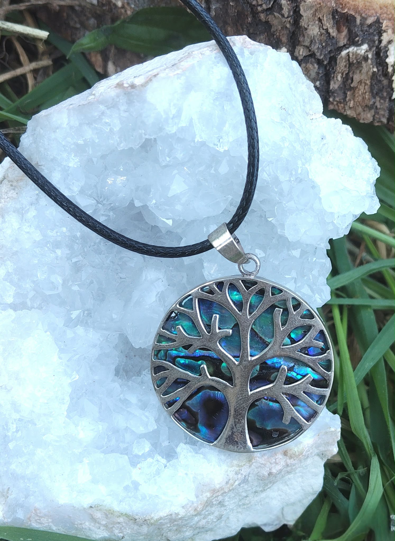 Pendentif arbre de vie en abalone et argent 925 sur cordon noir, posé sur amas clair de quartz près d’herbe et d’écorce, bijou en nacre naturelle lié à l’équilibre intérieur, au calme du cœur et au recentrage.