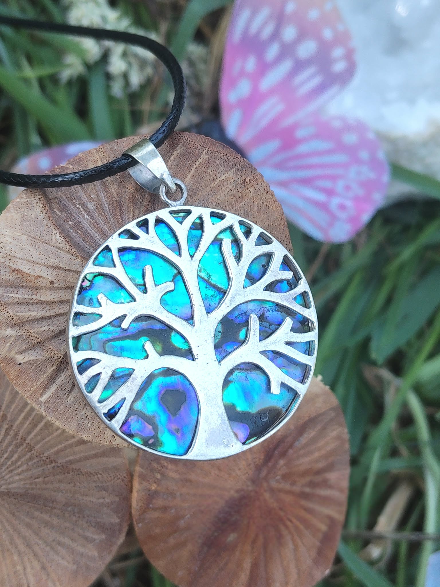 Pendentif arbre de vie en abalone et argent 925 sur cordon noir, posé sur rondin de bois avec herbes vertes et papillon rose flou, bijou en nacre naturelle évoquant la confiance douce, la paix intérieure et la sensibilité apaisée.
