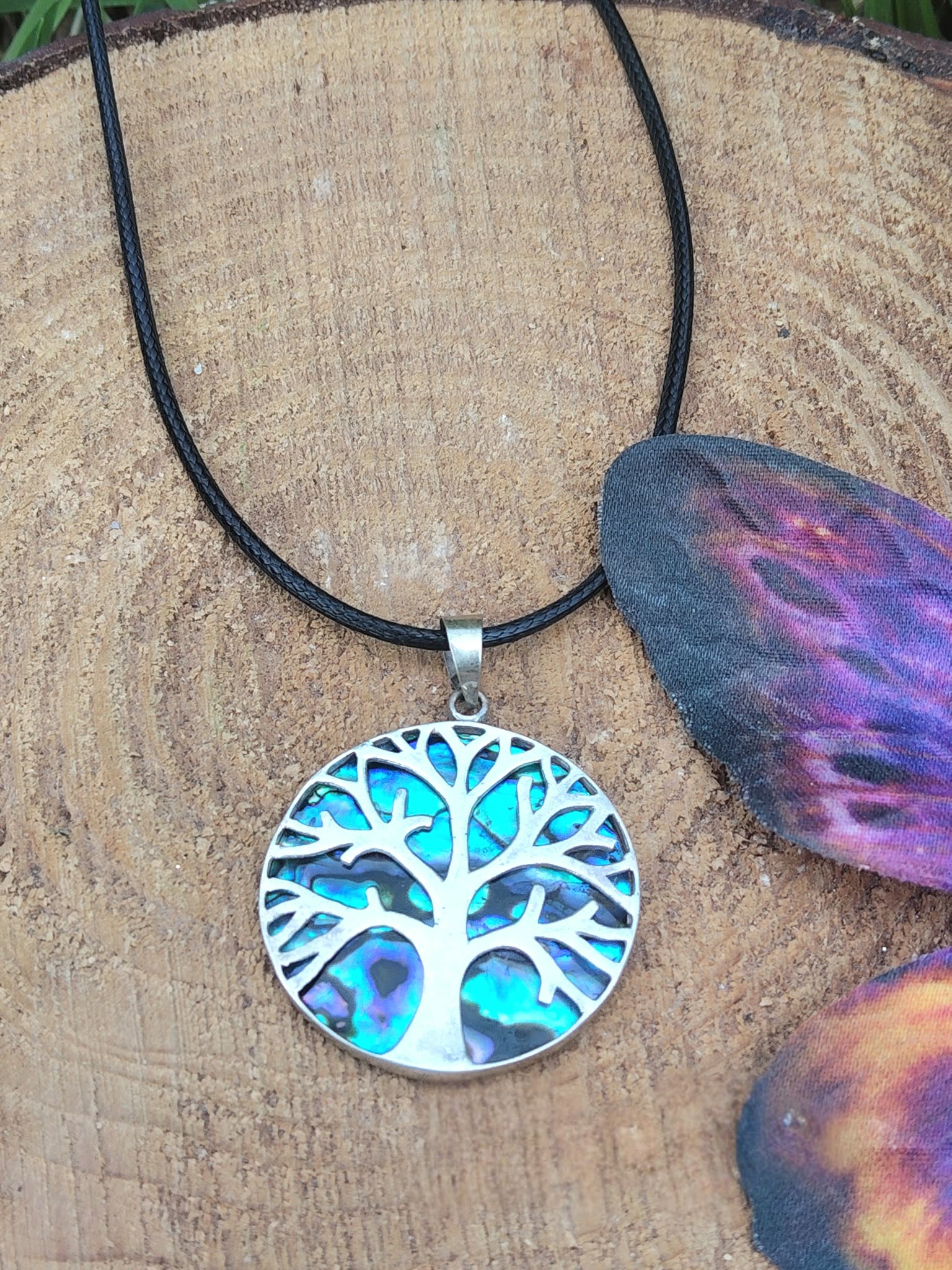 Pendentif arbre de vie en abalone et argent 925 sur cordon noir, posé sur rondin de bois avec papillon violet en arrière-plan, bijou en nacre naturelle évoquant l’expression sincère, la sérénité et l’harmonie intérieure.