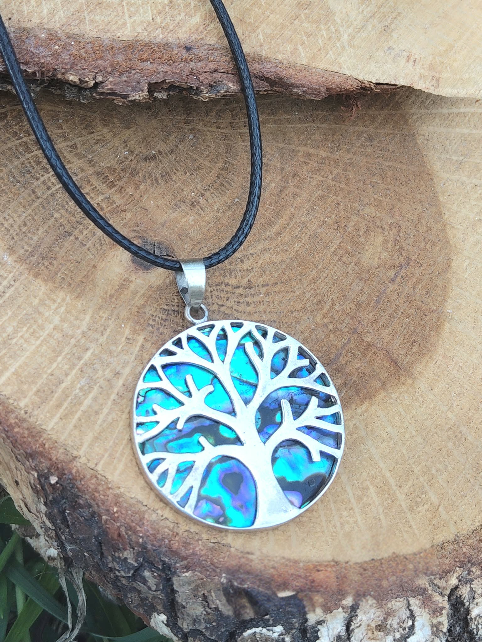 Pendentif arbre de vie en abalone et argent 925 sur cordon noir, posé sur tranche de bois près d’un papillon peint, bijou en nacre naturelle associé à l’apaisement, à la présence à soi et à la confiance intérieure.