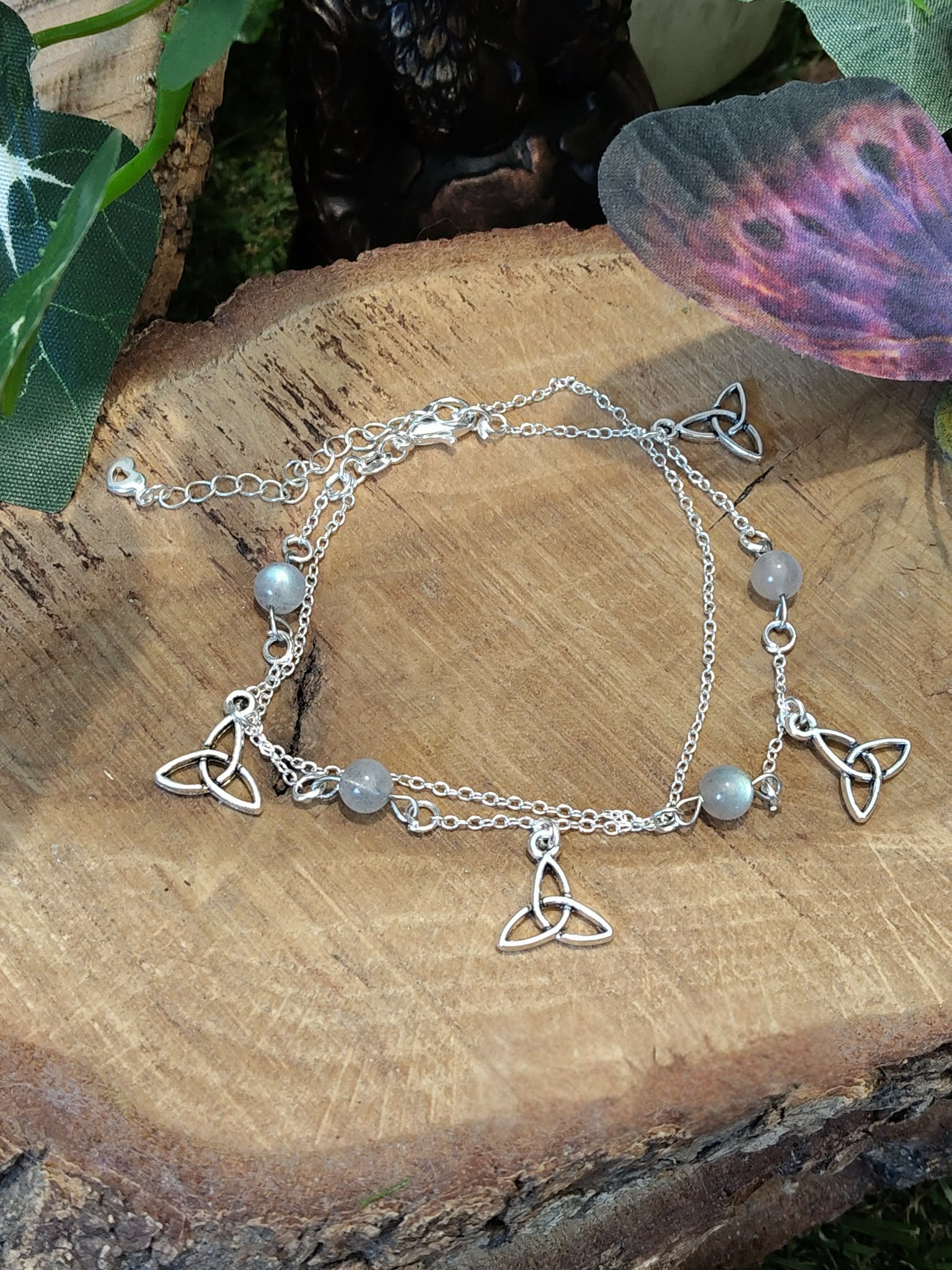 Chaîne de cheville en acier inoxydable avec perles de labradorite et breloques triquetra, posée sur rondin de bois près d’un papillon décoratif, idéale pour l’ancrage et la protection au quotidien.