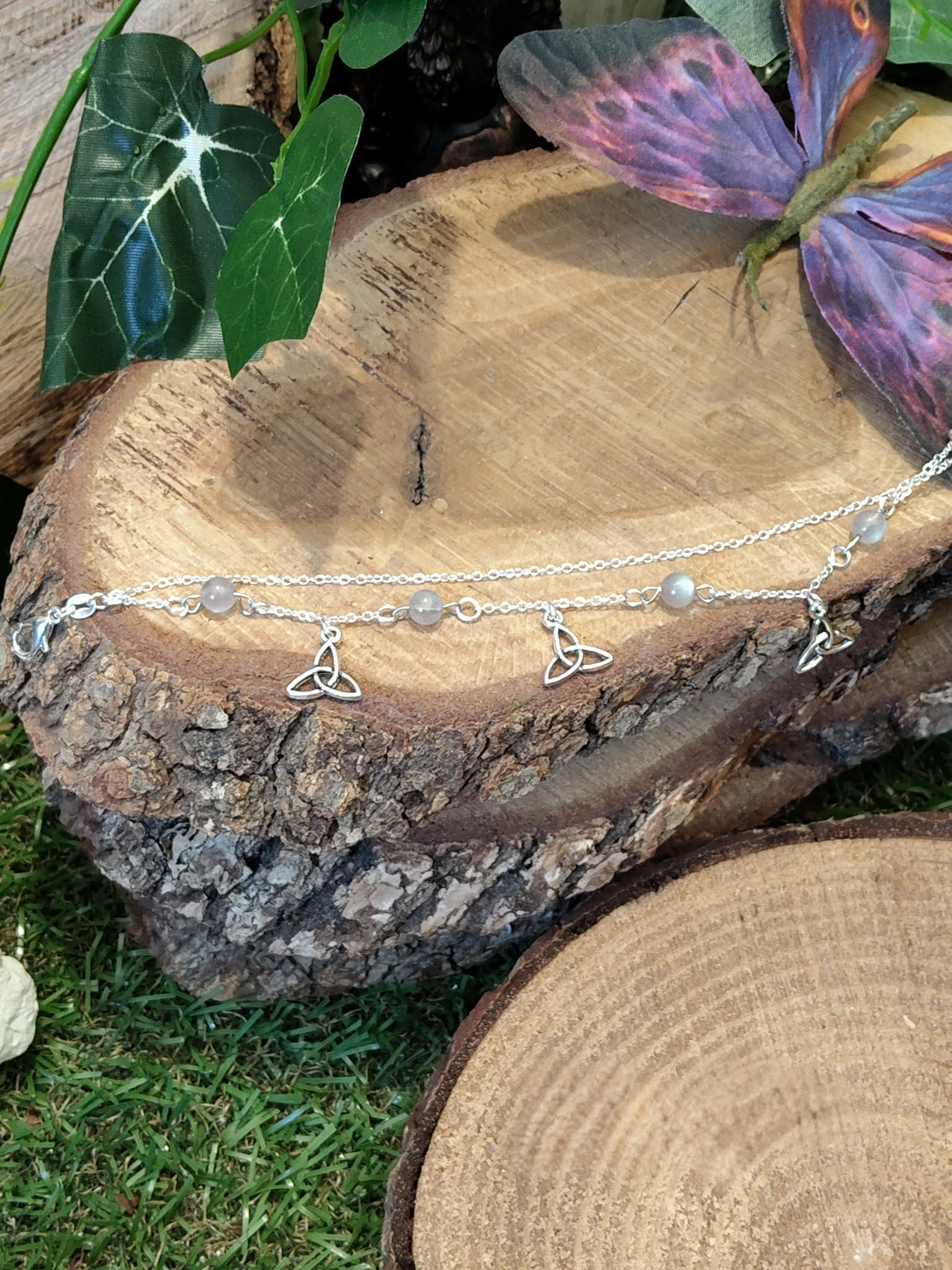 Chaîne de cheville création Magic Gaïa en acier inoxydable, breloques triquetra et perles de labradorite, posée sur rondin de bois avec lierre et papillon violet, pour protection et confiance.