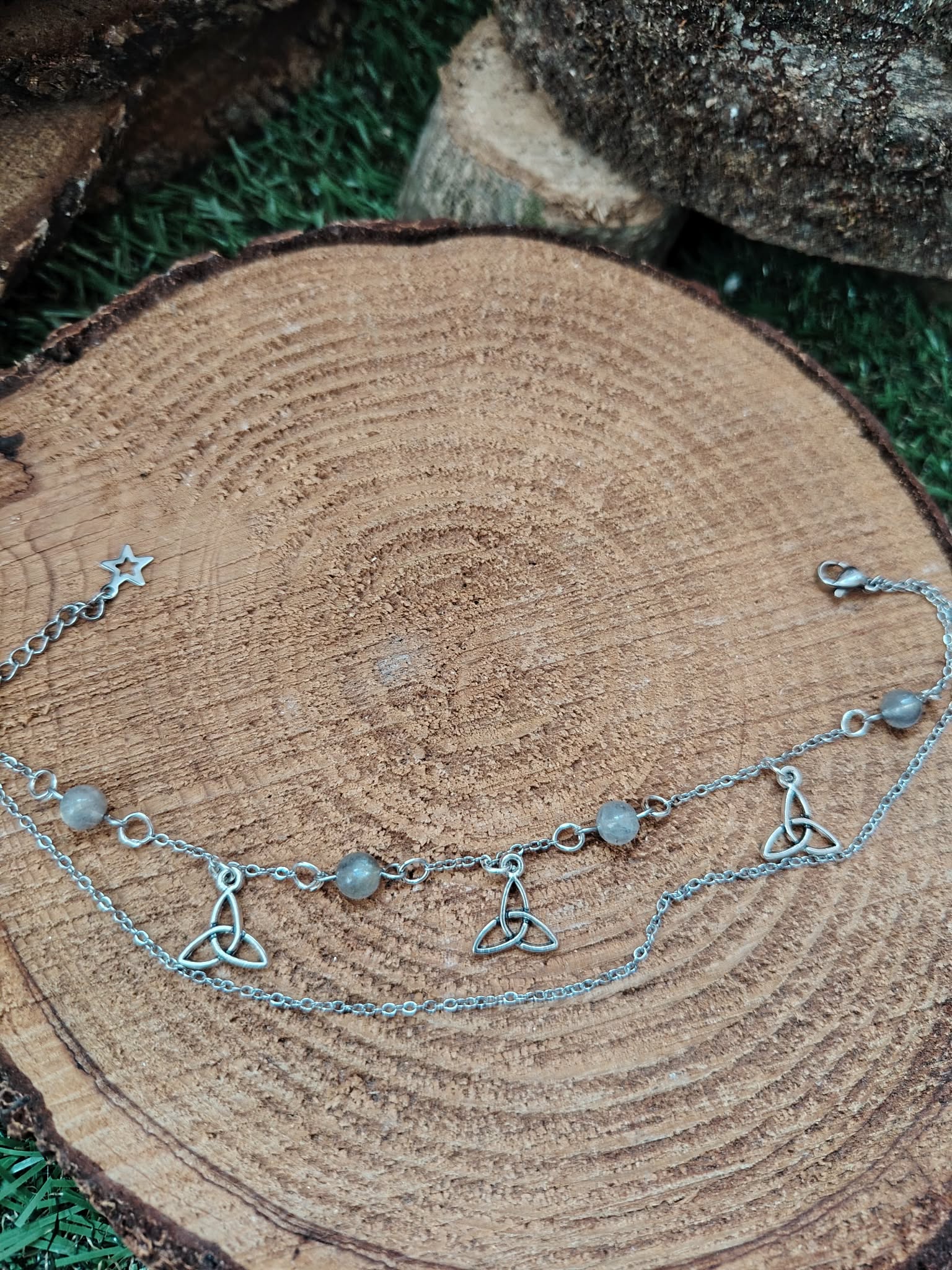 Chaîne de cheville en acier inoxydable avec perles de labradorite et breloques triquetra, posée sur un rondin de bois aux cernes visibles, pour l’ancrage, la protection et la confiance