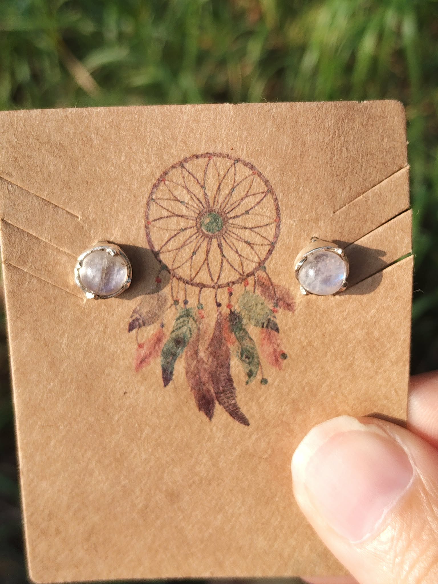 Boucles d’oreilles en argent 925 serties de péristérite sur carte kraft attrape-rêves tenue entre les doigts, fond végétal flou, pour apaiser et clarifier l’esprit.